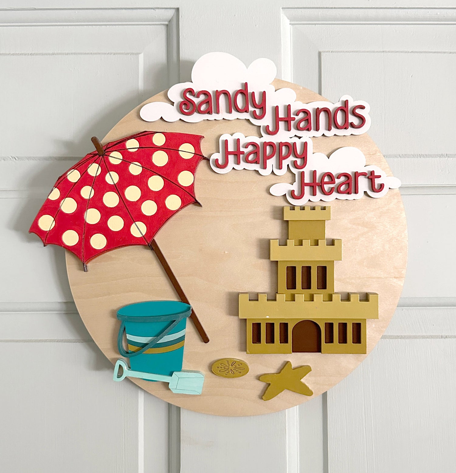 A laser cut round door hangerwith an umbrella, pail and bucket, sand castle, starfish, sanddollar, clouds and the words Sandy Hands Happy Heart.