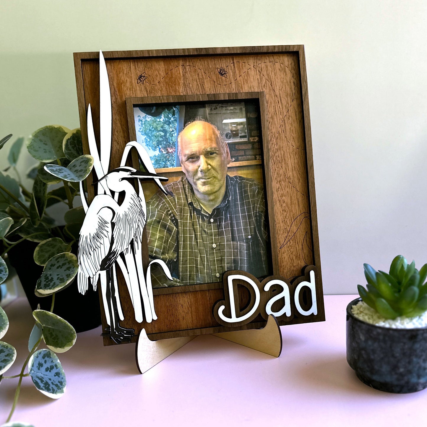 Wooden photo frame with a picture of a man, decorative angel wings, and 'Dad' text on a white surface with plants.