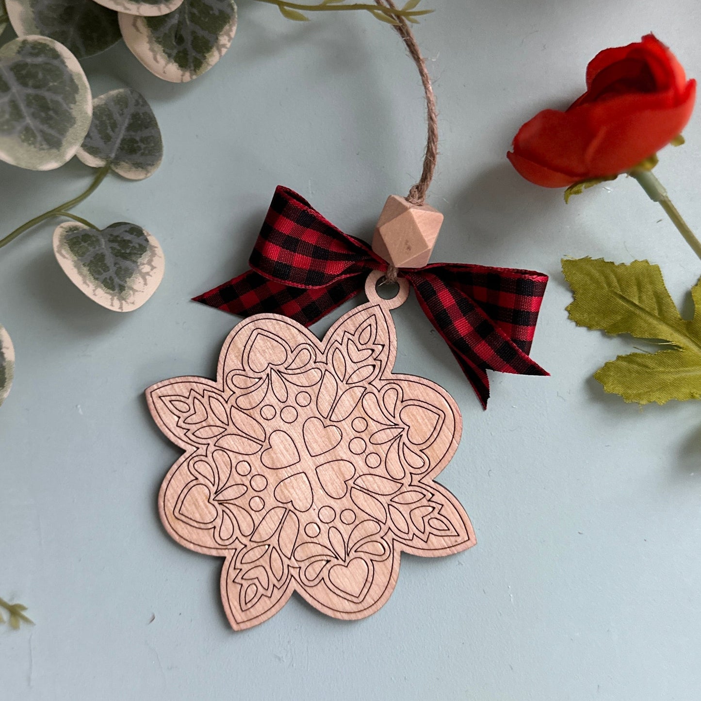 Decorative copper ornament with a red and black checkered bow, surrounded by greenery and flowers on a light gray background.