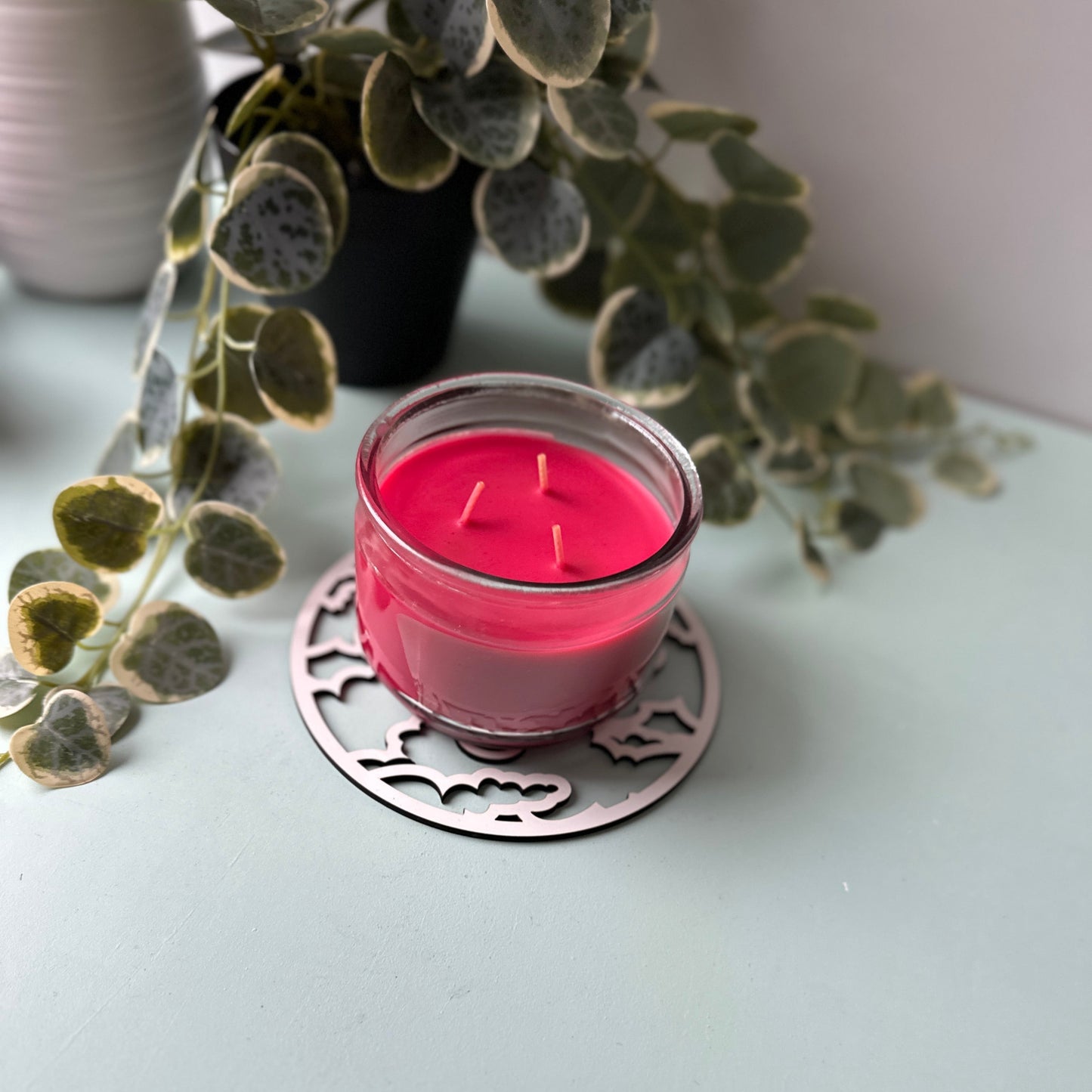 Pink candle in a glass holder on a decorative coaster with greenery in the background.