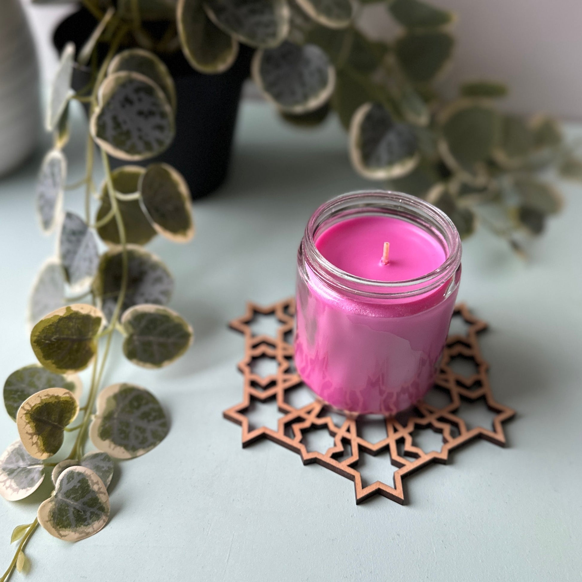 Pink candle in a glass holder on a decorative trivet with a plant in the background.