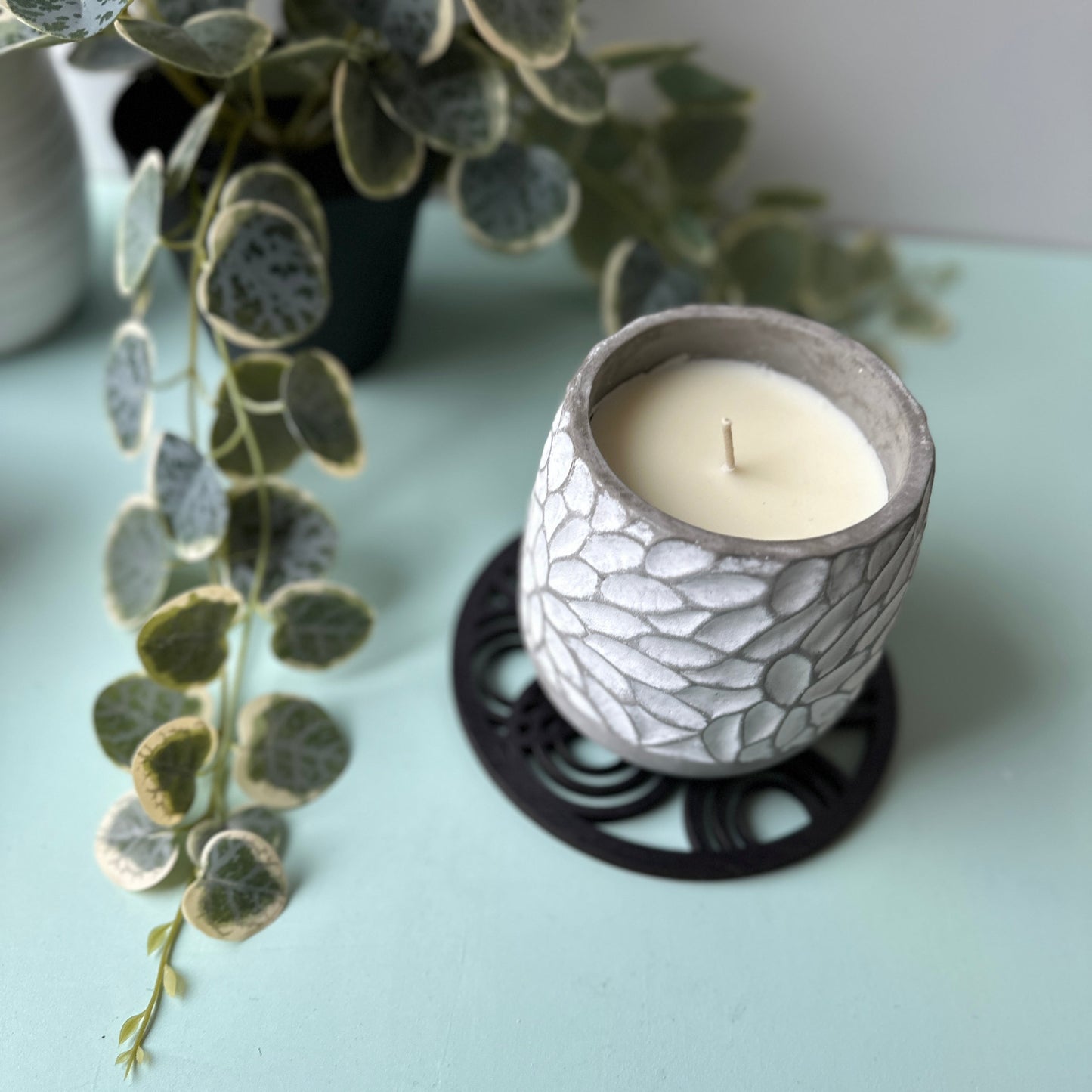 Ceramic candle holder with a white candle on a light blue surface, accompanied by a plant.