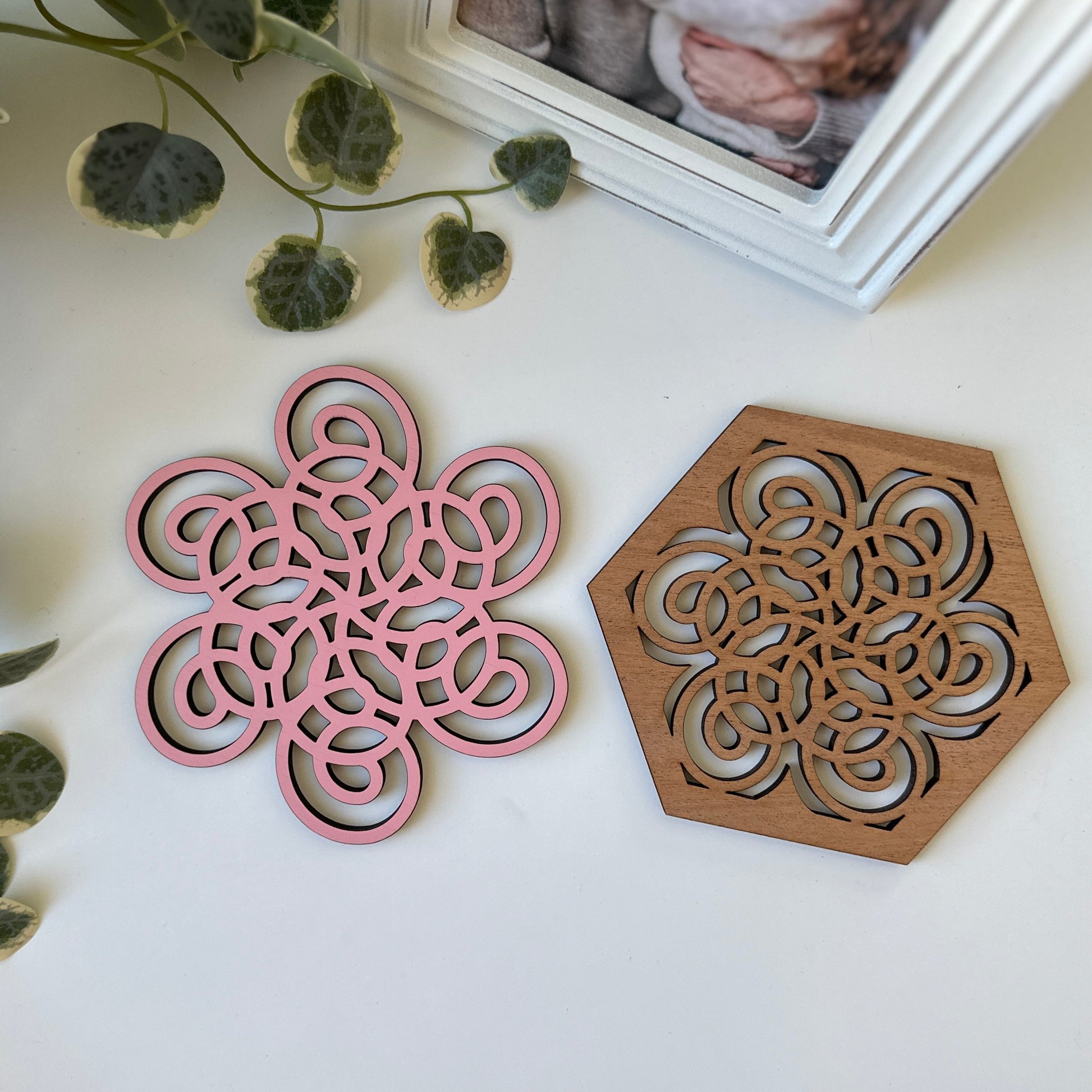 Pink and wooden geometric coasters on a white surface with green leaves.