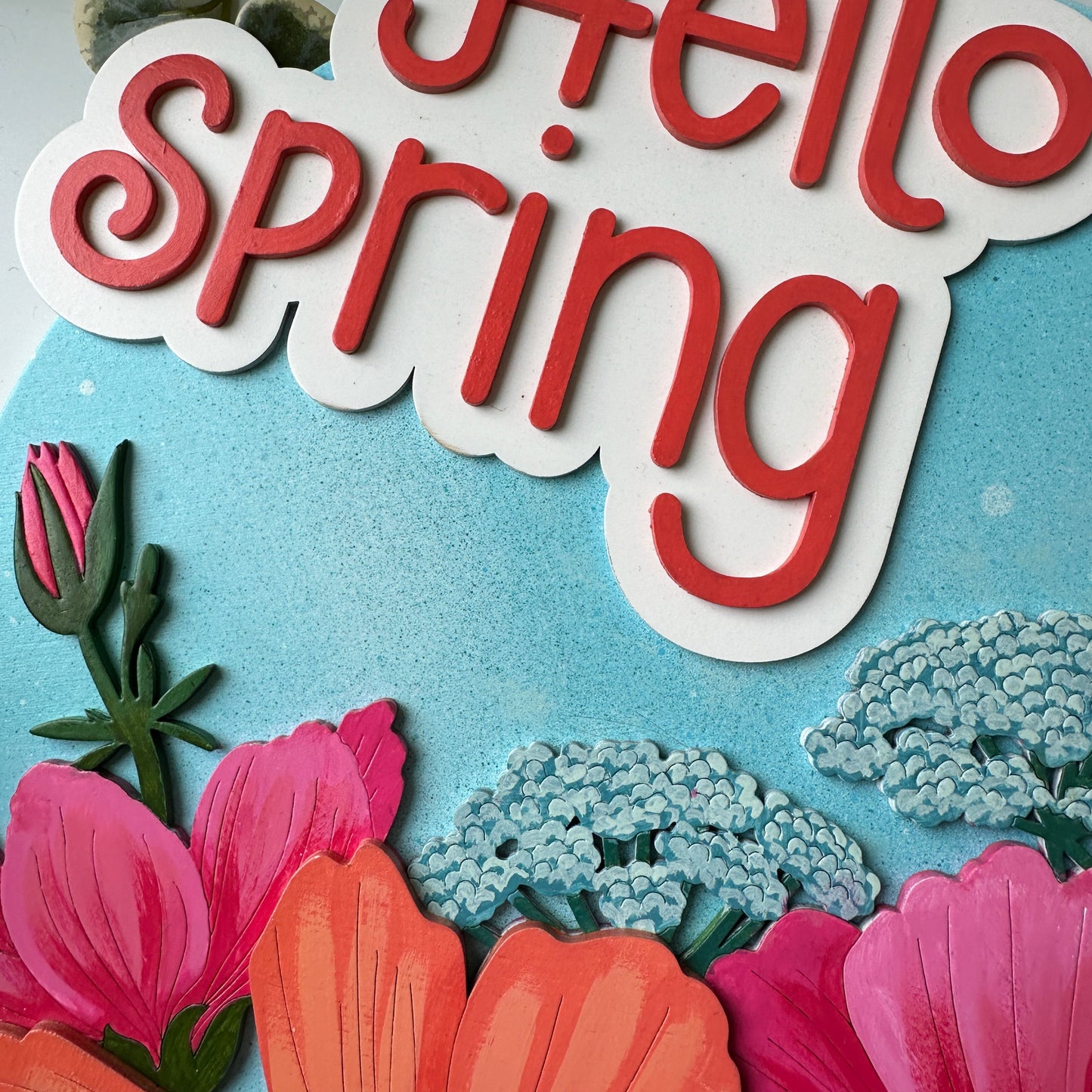 Decorative sign with 'Hello Spring' text surrounded by colorful flowers on a light blue background