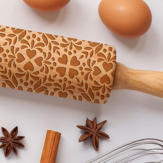 Wooden rolling pin with heart pattern on a white surface with eggs, star anise, cinnamon stick, and whisk.