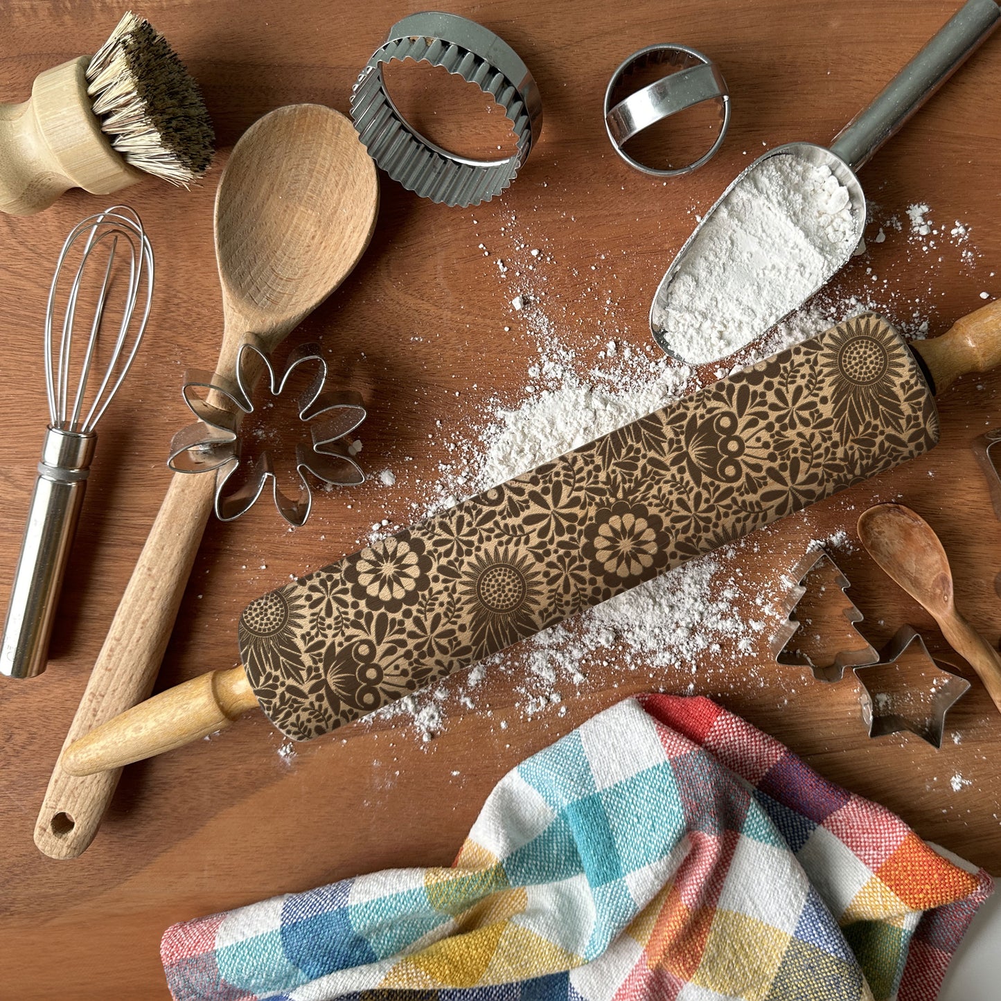 Baking tools including a rolling pin with embossed designs, wooden spoon, whisk, and cookie cutters on a wooden surface.
