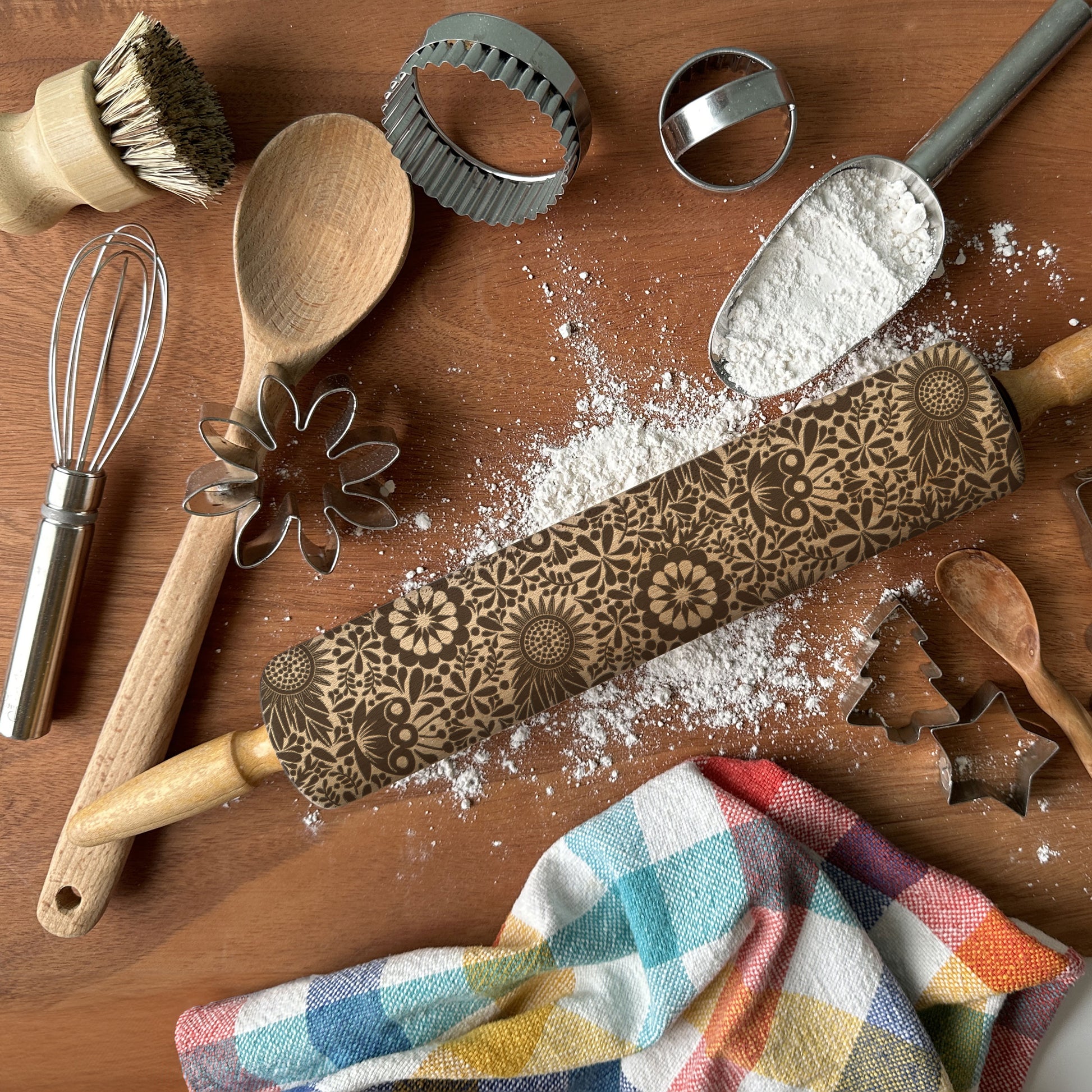 Baking tools including a rolling pin with embossed designs, wooden spoon, whisk, and cookie cutters on a wooden surface.