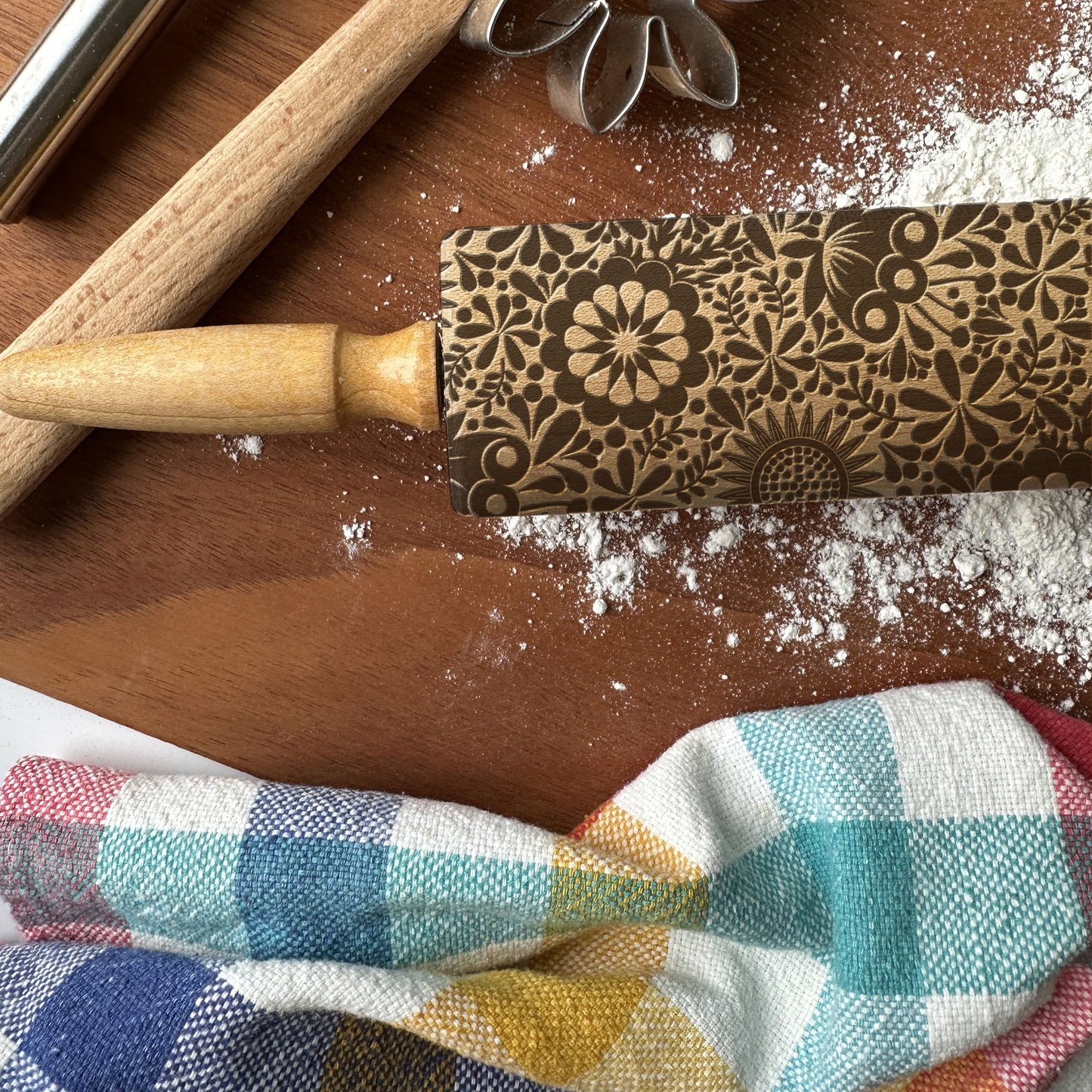 Decorative rolling pin with floral patterns on a wooden surface with flour and a checkered cloth.