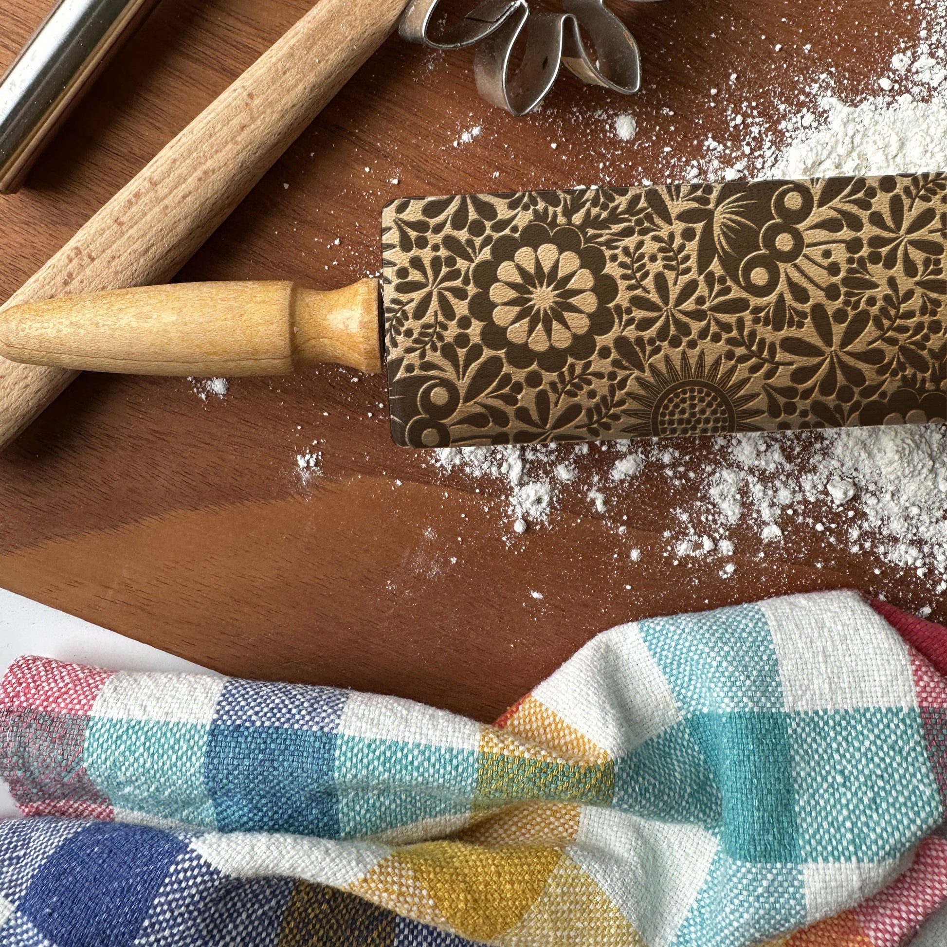Decorative rolling pin with floral patterns on a wooden surface with flour and a checkered cloth.