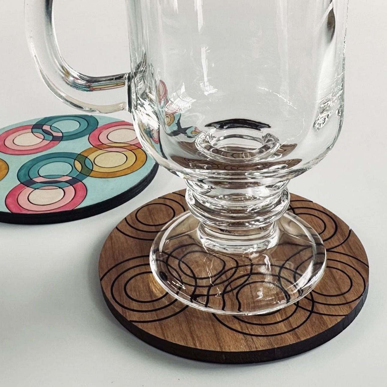 Glass mug on a wooden coaster with a colorful circular design, on a light gray background
