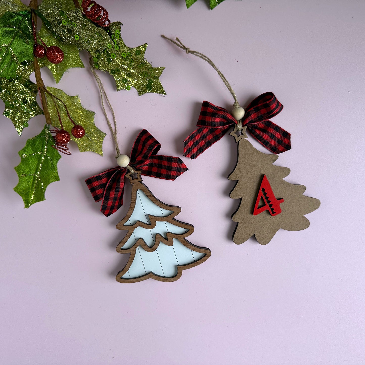 Laser-cut layered Christmas tree ornaments with buffalo plaid ribbon bows, hanging against a holiday backdrop