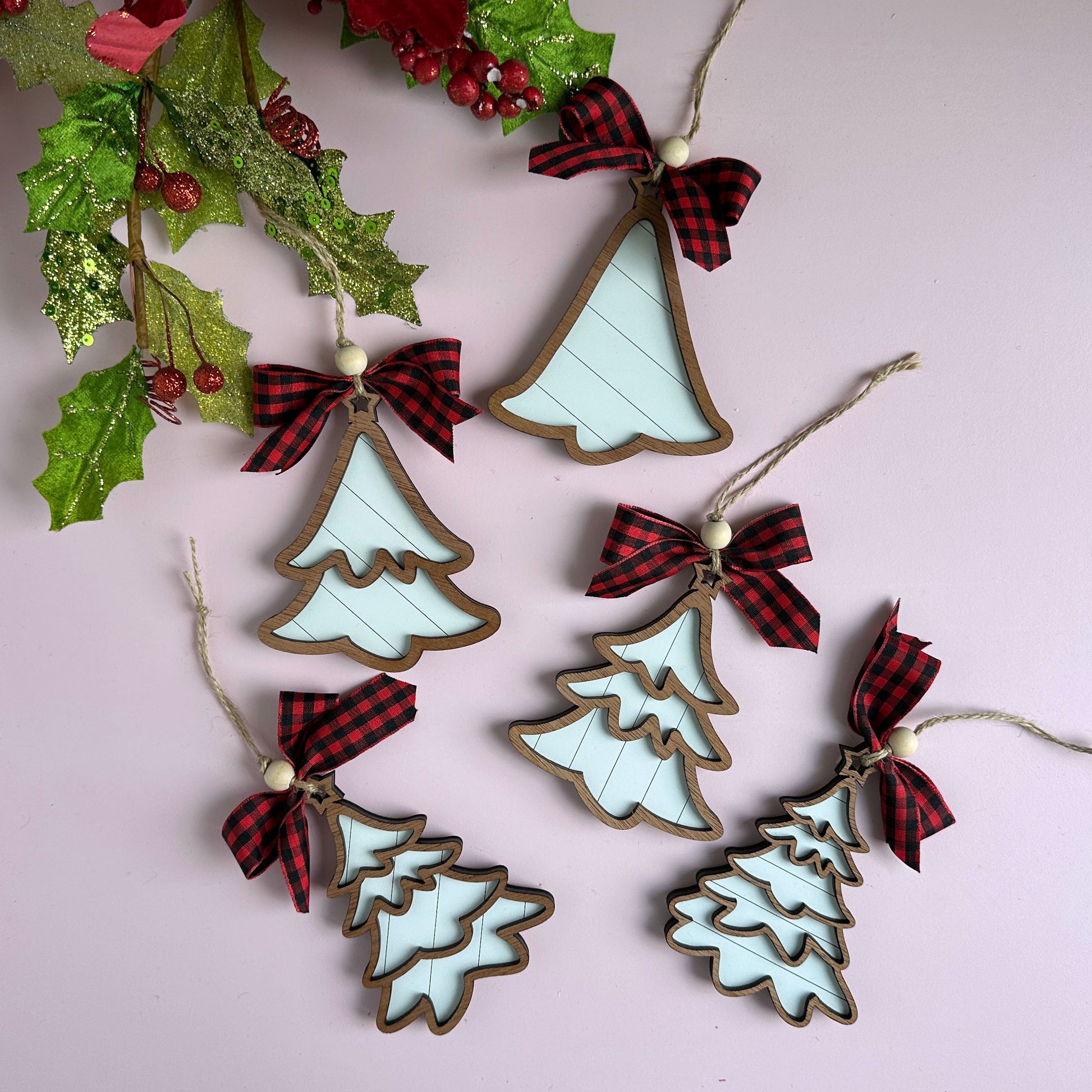 Laser-cut layered Christmas tree ornaments with buffalo plaid ribbon bows, hanging against a holiday backdrop