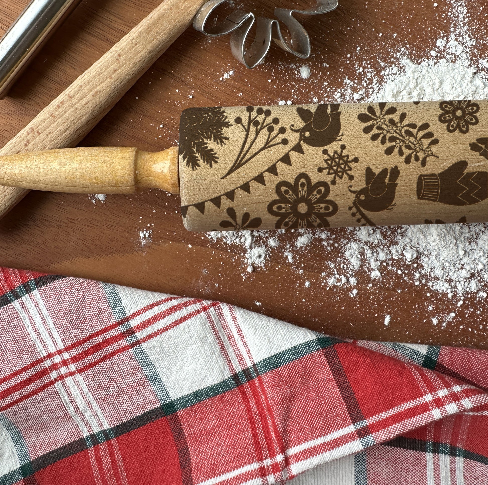 Decorative rolling pin with floral and bird patterns on a wooden surface with flour and a plaid cloth.