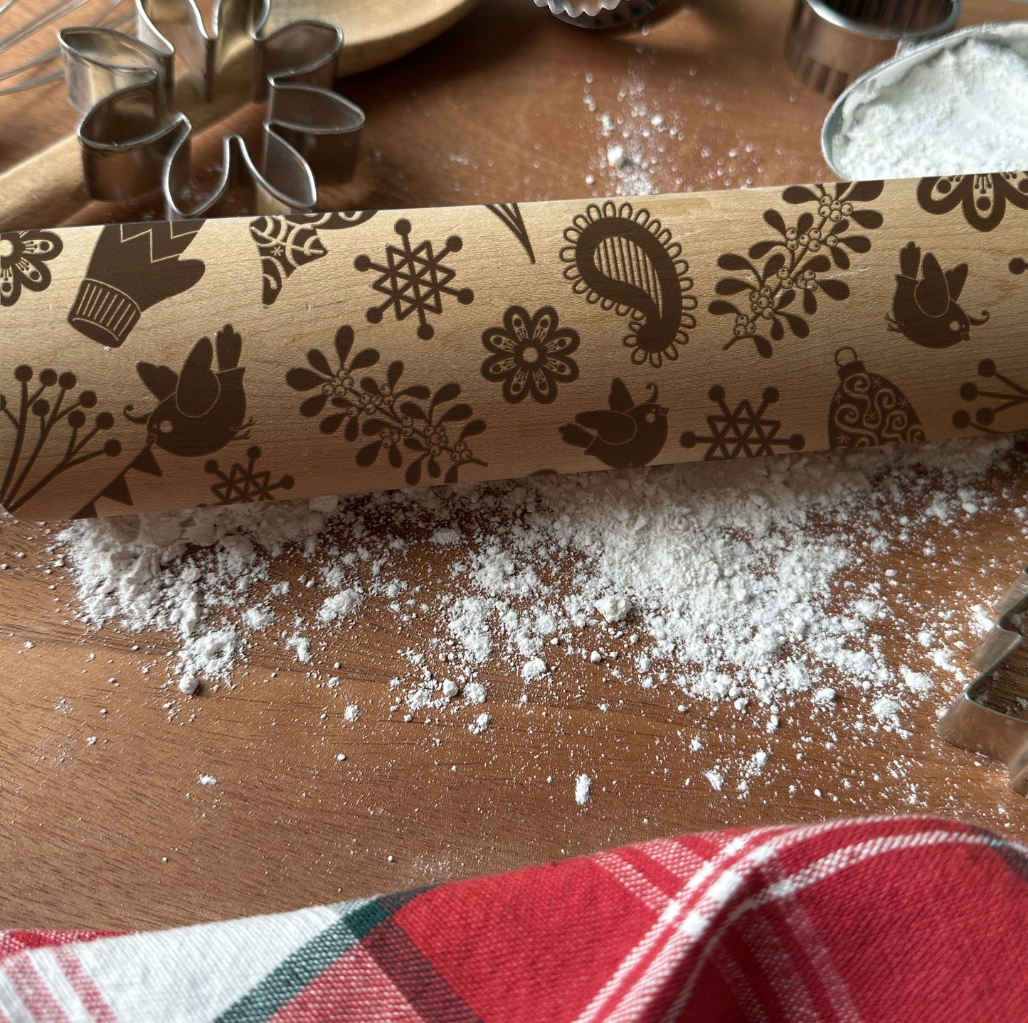 Wooden rolling pin with embossed patterns on a wooden surface with flour and a red checkered towel.