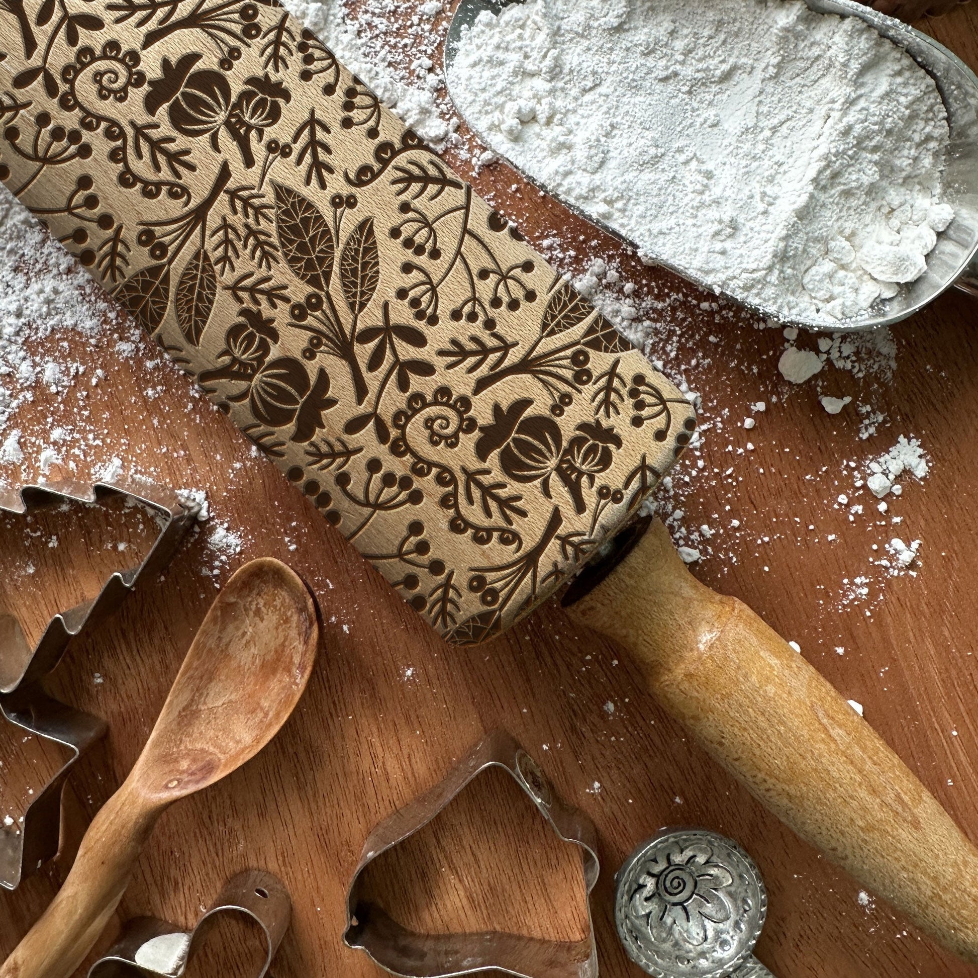 Decorative rolling pin with floral patterns on a wooden surface with flour and cookie cutters.