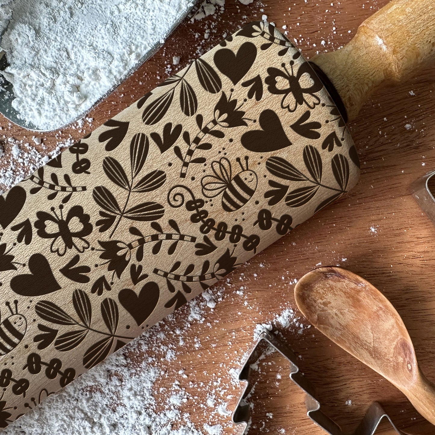Wooden rolling pin with floral patterns on a wooden surface with flour and cookie cutters.