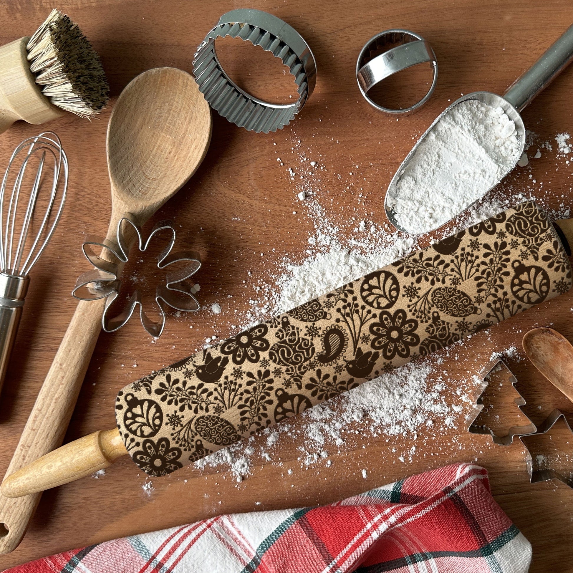 Baking tools including a rolling pin with embossed designs, wooden spoon, whisk, and cookie cutters on a wooden surface.