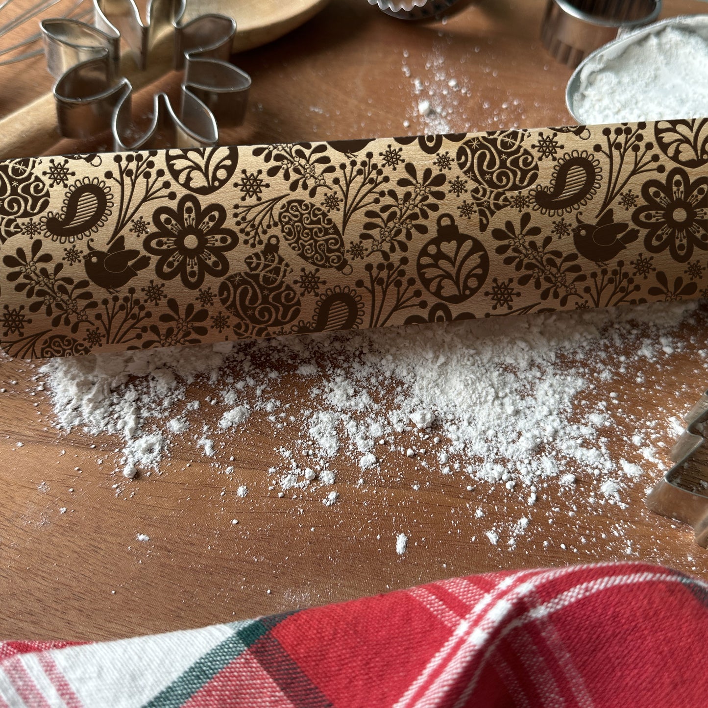 Decorative rolling pin with floral patterns on a wooden surface with flour and a red checkered towel.