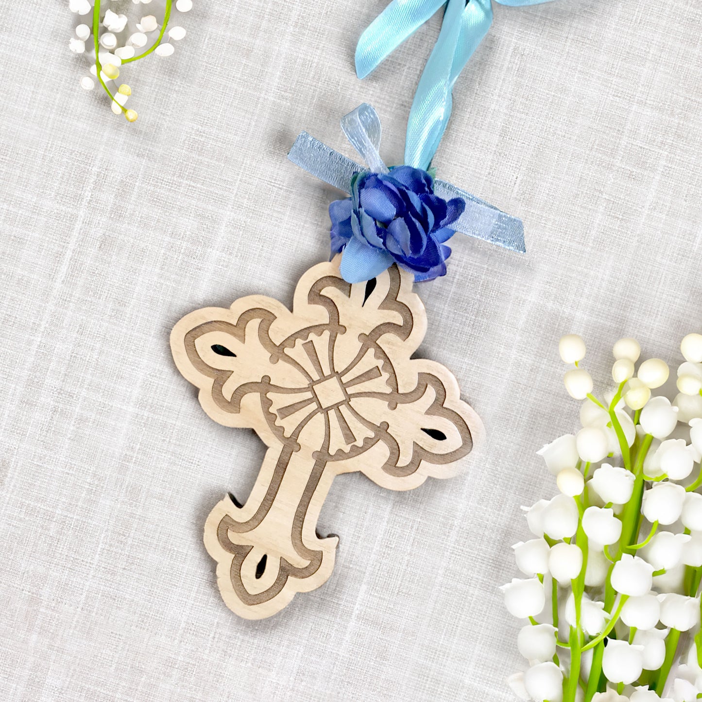Wooden cross ornament with blue flower and ribbon on a textured surface with flowers.