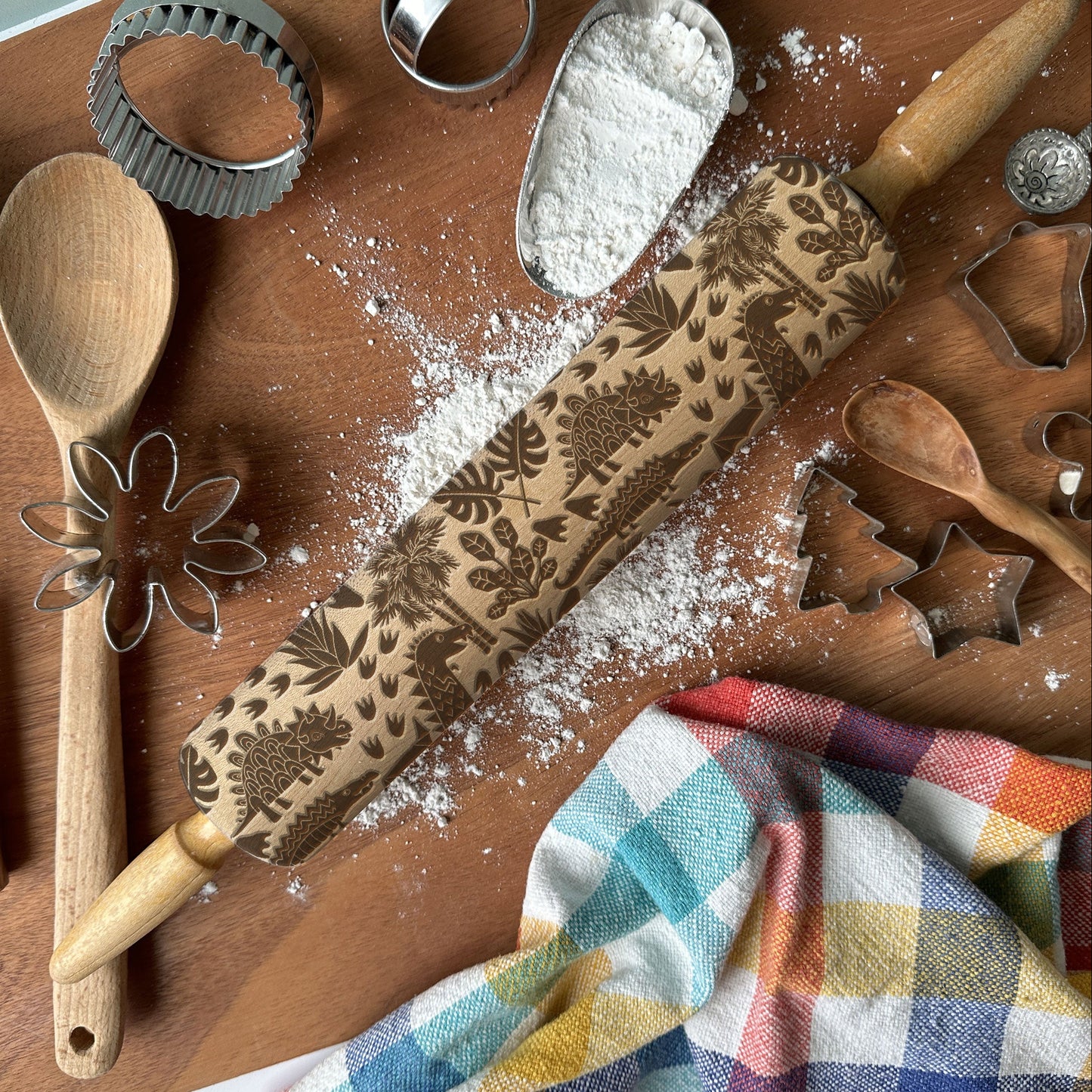 Baking tools including a rolling pin with floral patterns, wooden spoon, whisk, and flour on a wooden surface.
