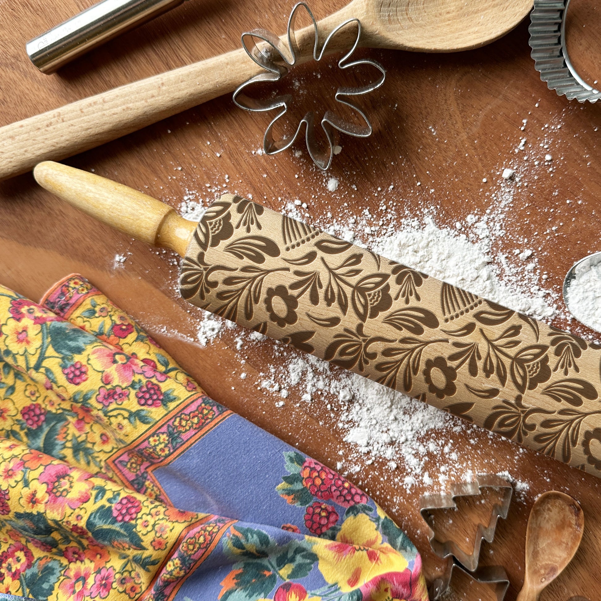 Decorative rolling pin with floral patterns on a wooden surface with flour and kitchen tools.