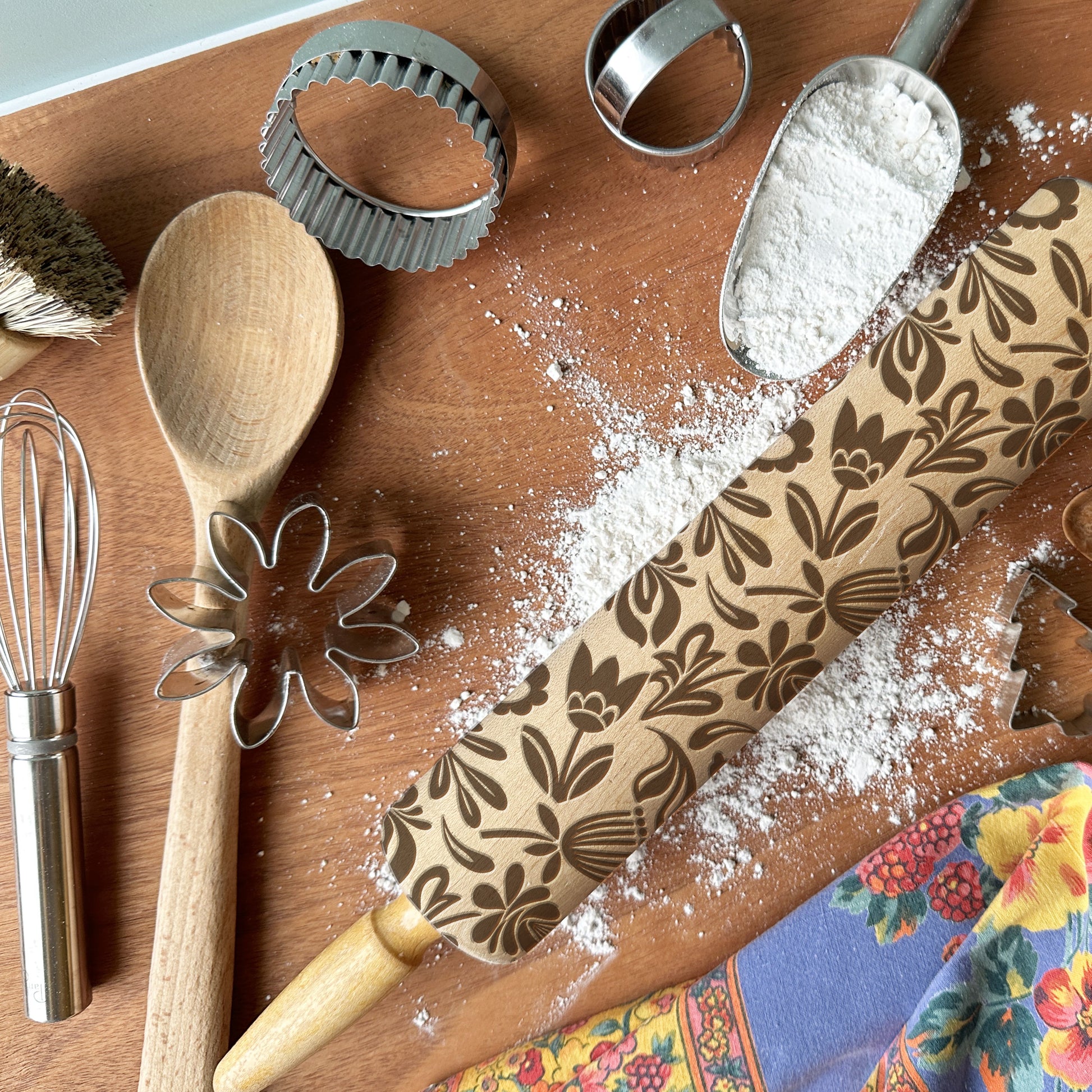 Baking tools including a wooden spoon, rolling pin with floral patterns, and cookie cutters on a wooden surface.