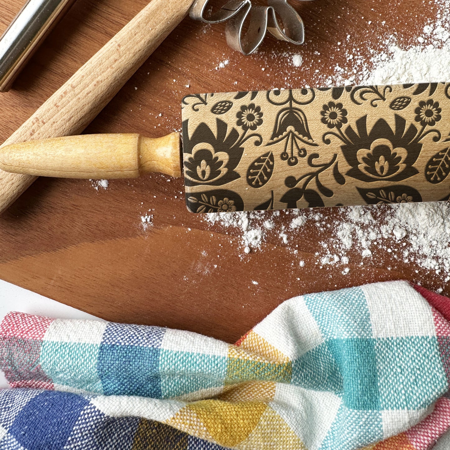 Decorative rolling pin with floral patterns on a wooden surface with flour and a checkered cloth.