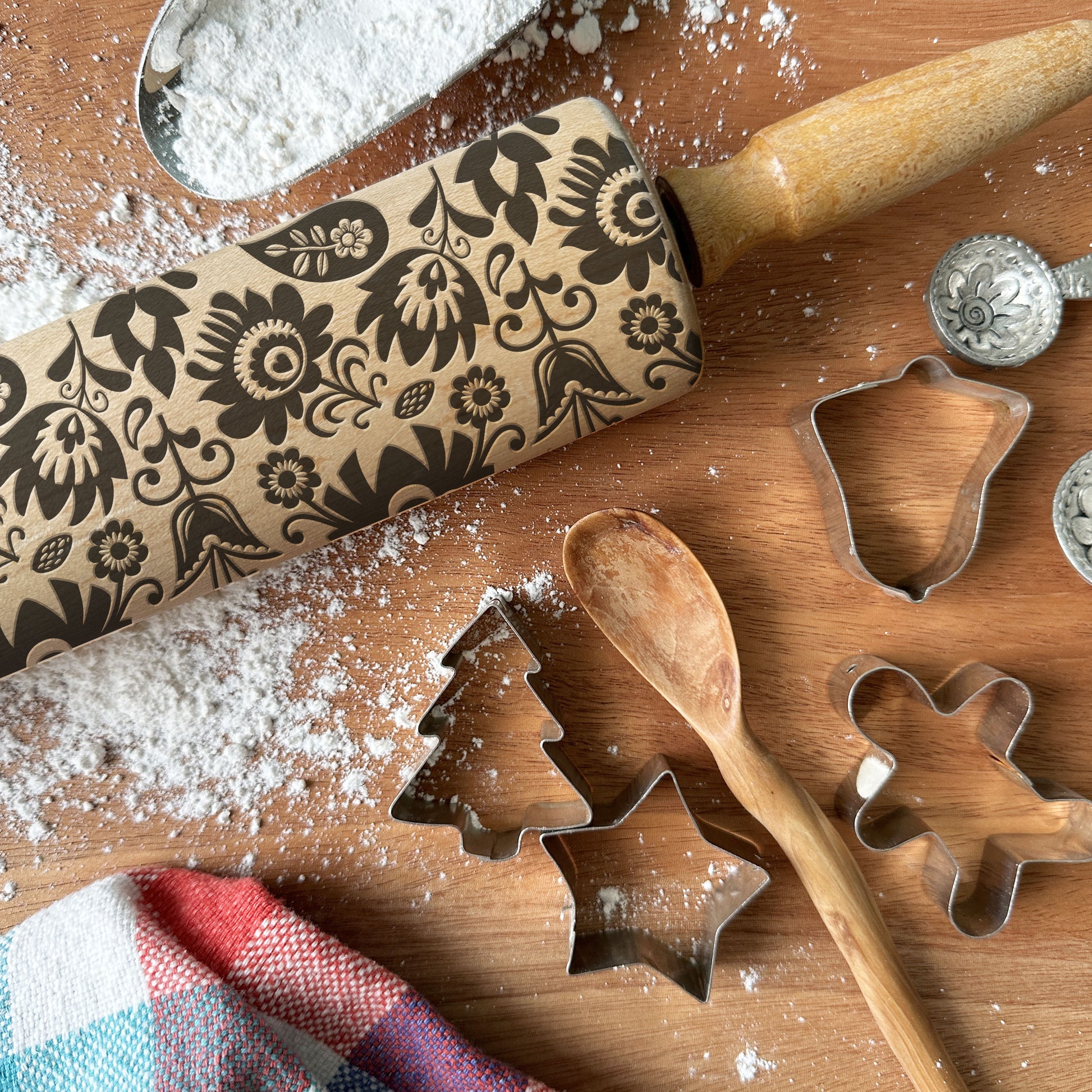 Decorative rolling pin with floral patterns on a wooden surface with cookie cutters and flour.