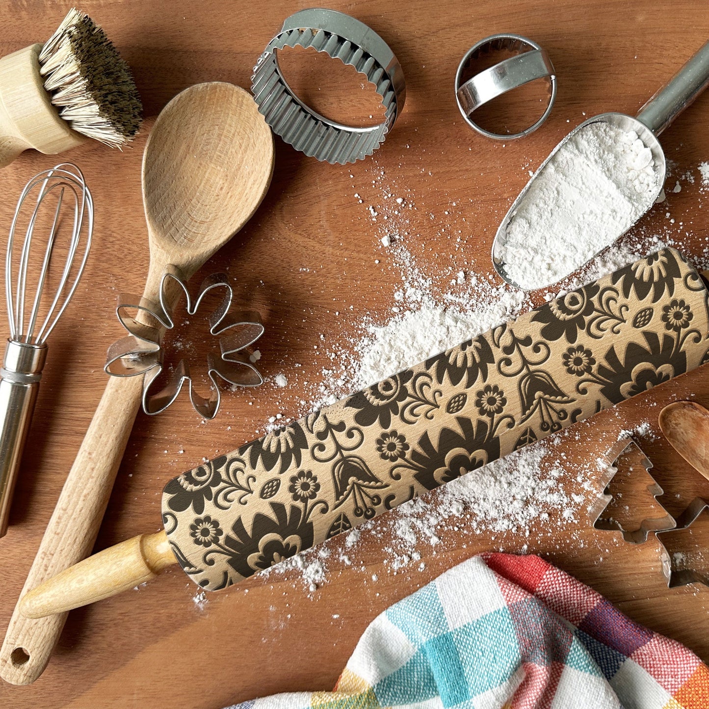 Baking tools including a rolling pin with decorative patterns, wooden spoon, whisk, and cookie cutters on a wooden surface.