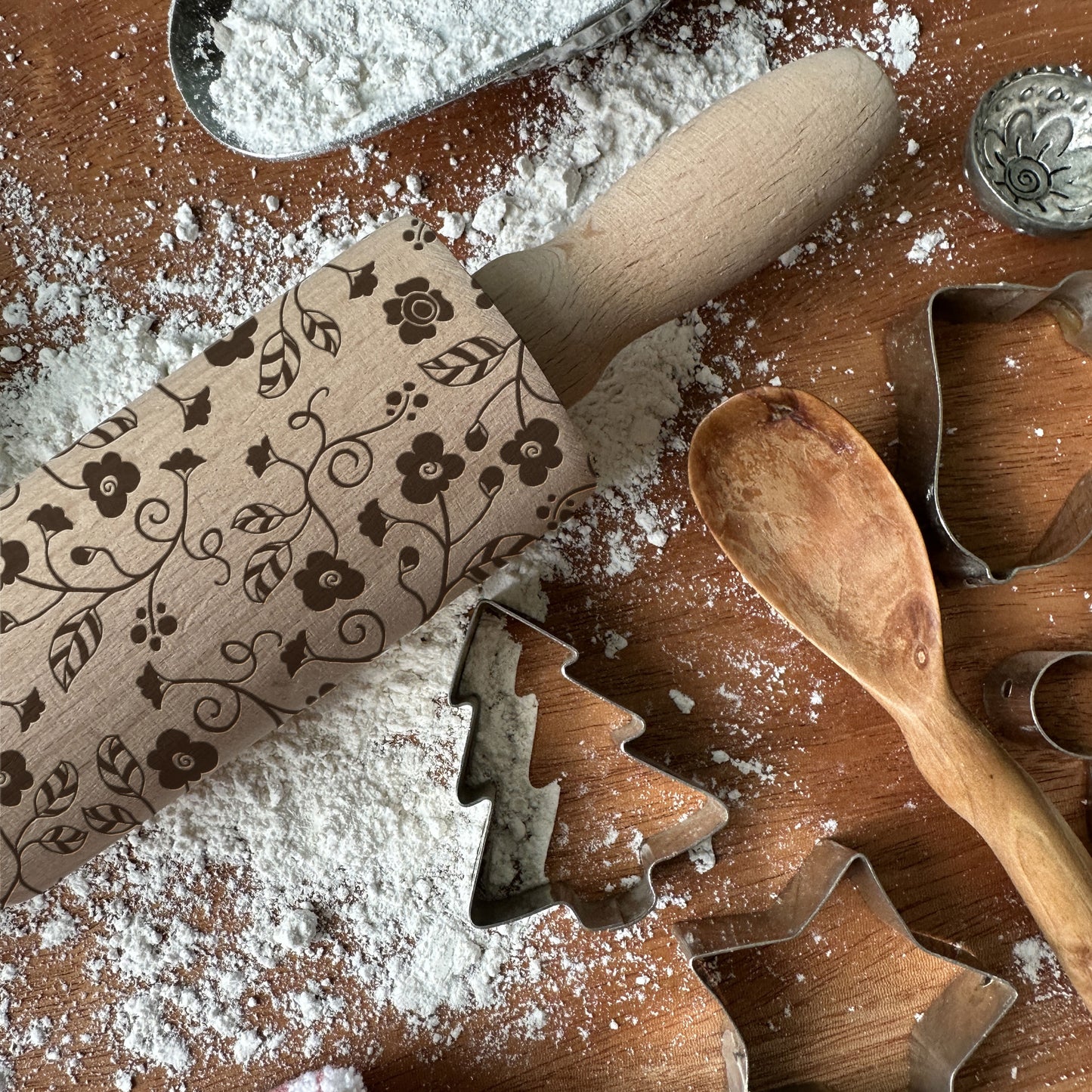 Decorative rolling pin with floral patterns on a flour-dusted surface with cookie cutters and a wooden spoon.