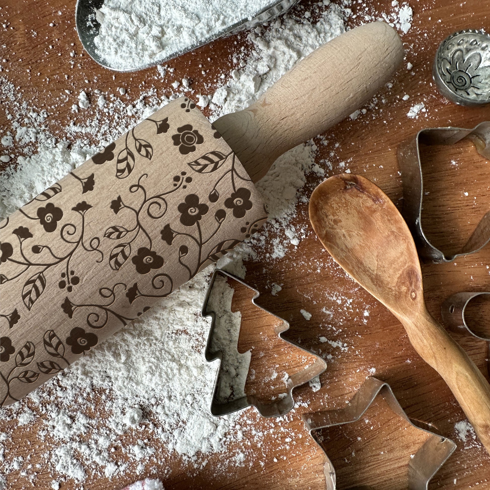 Decorative rolling pin with floral patterns on a flour-dusted surface with cookie cutters and a wooden spoon.