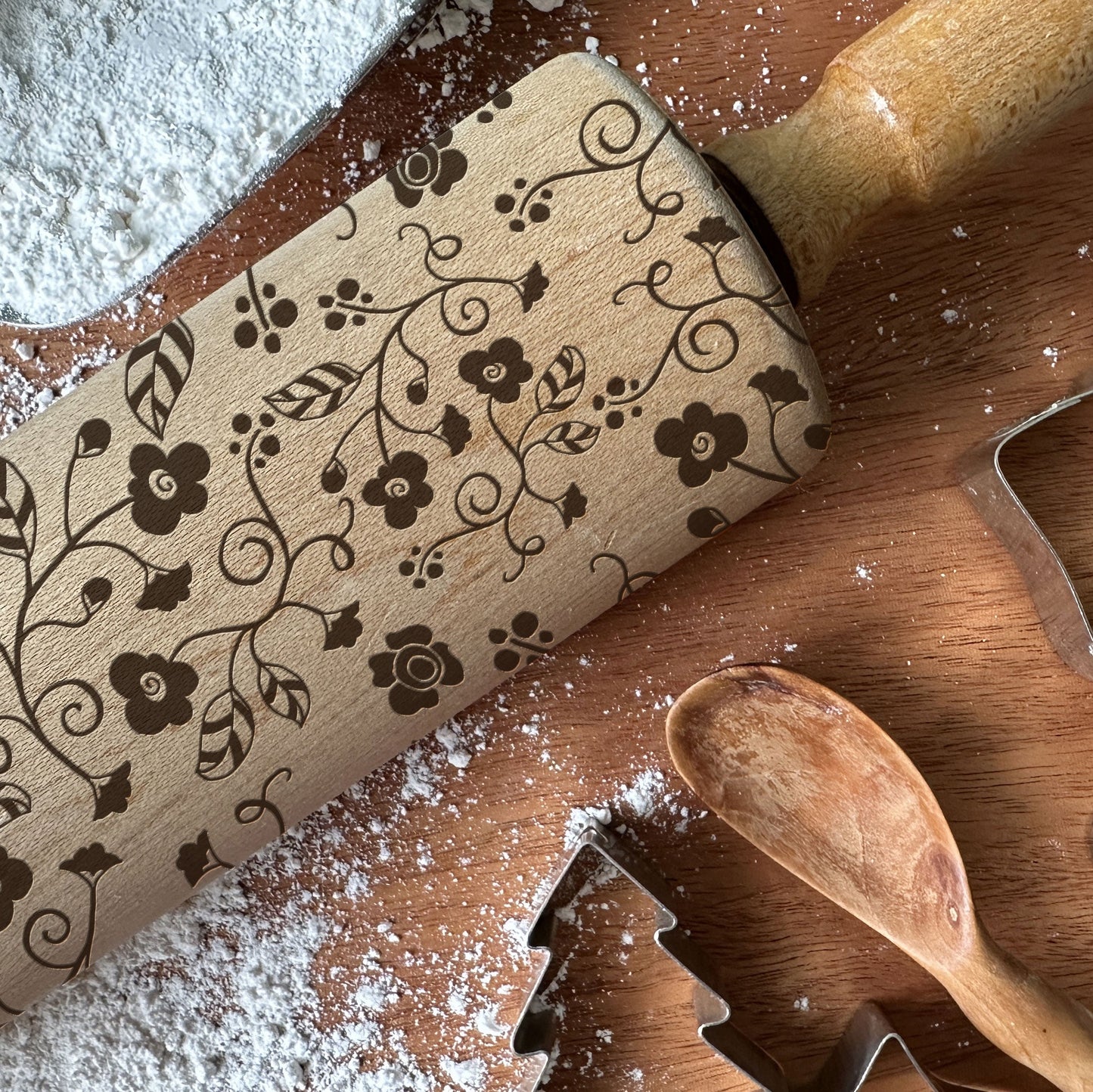 Wooden rolling pin with floral patterns on a wooden surface with flour and cookie cutters.