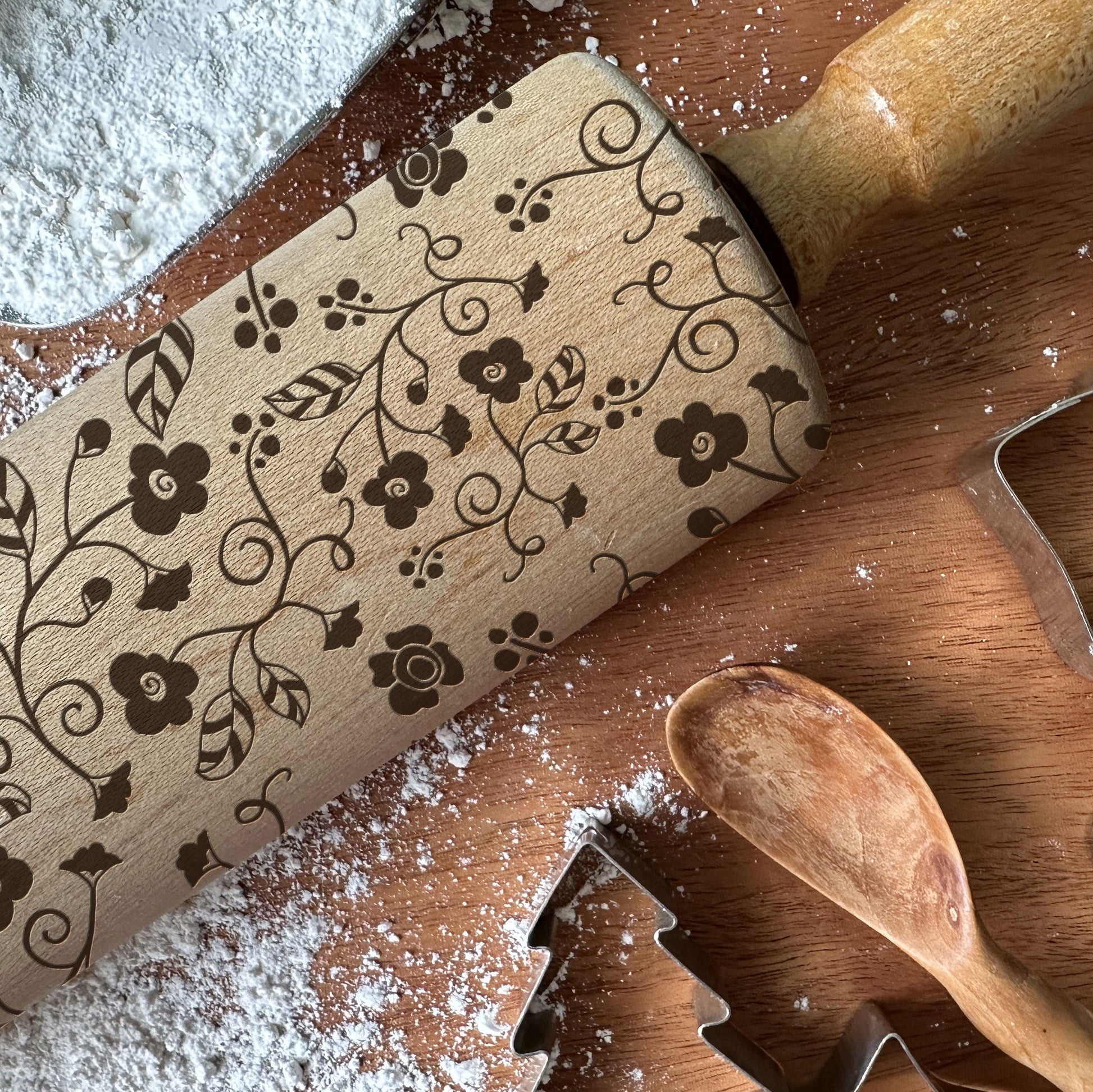 Wooden rolling pin with floral patterns on a wooden surface with flour and cookie cutters.