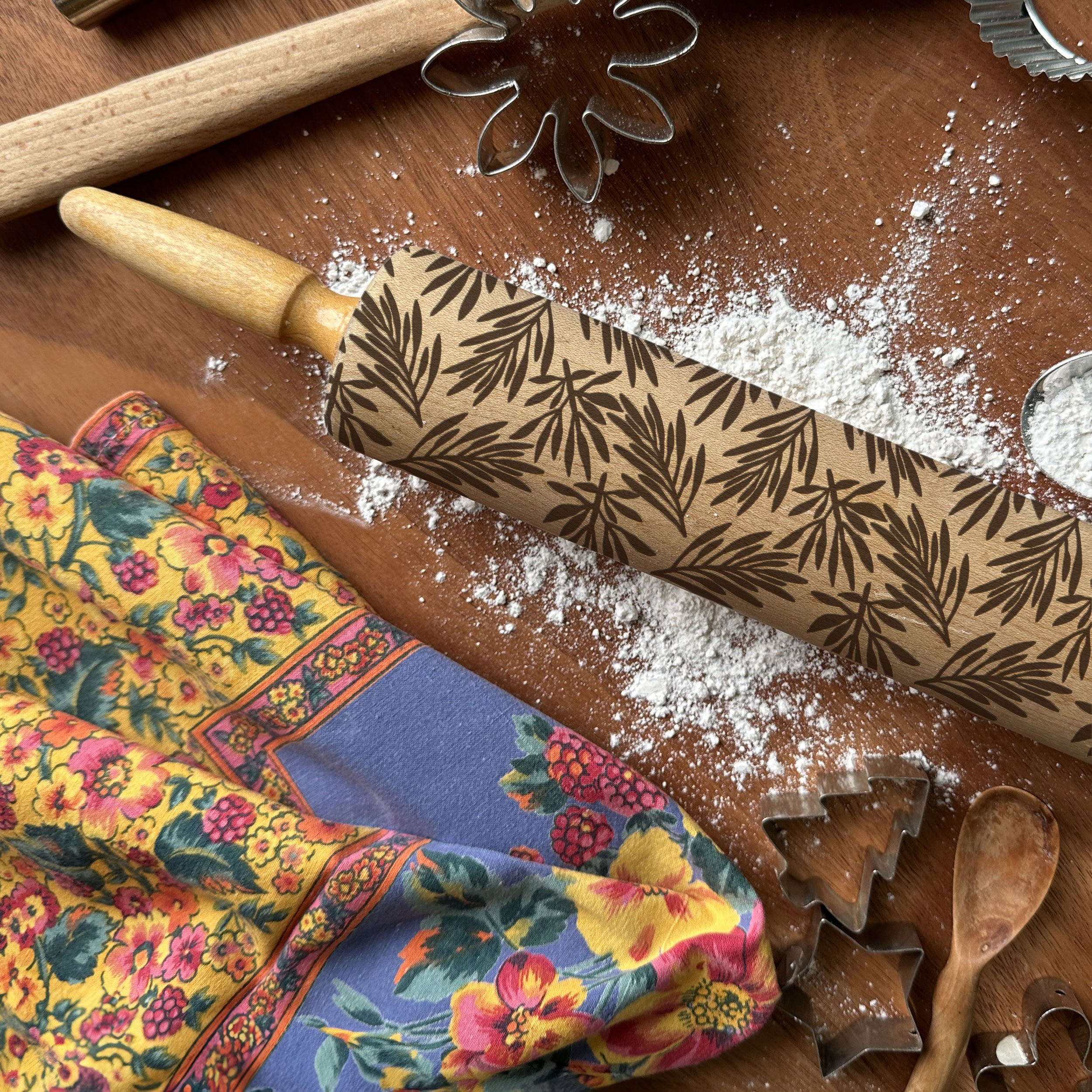 Wooden rolling pin with leaf pattern on a wooden surface with flour, colorful fabric, and cookie cutters.