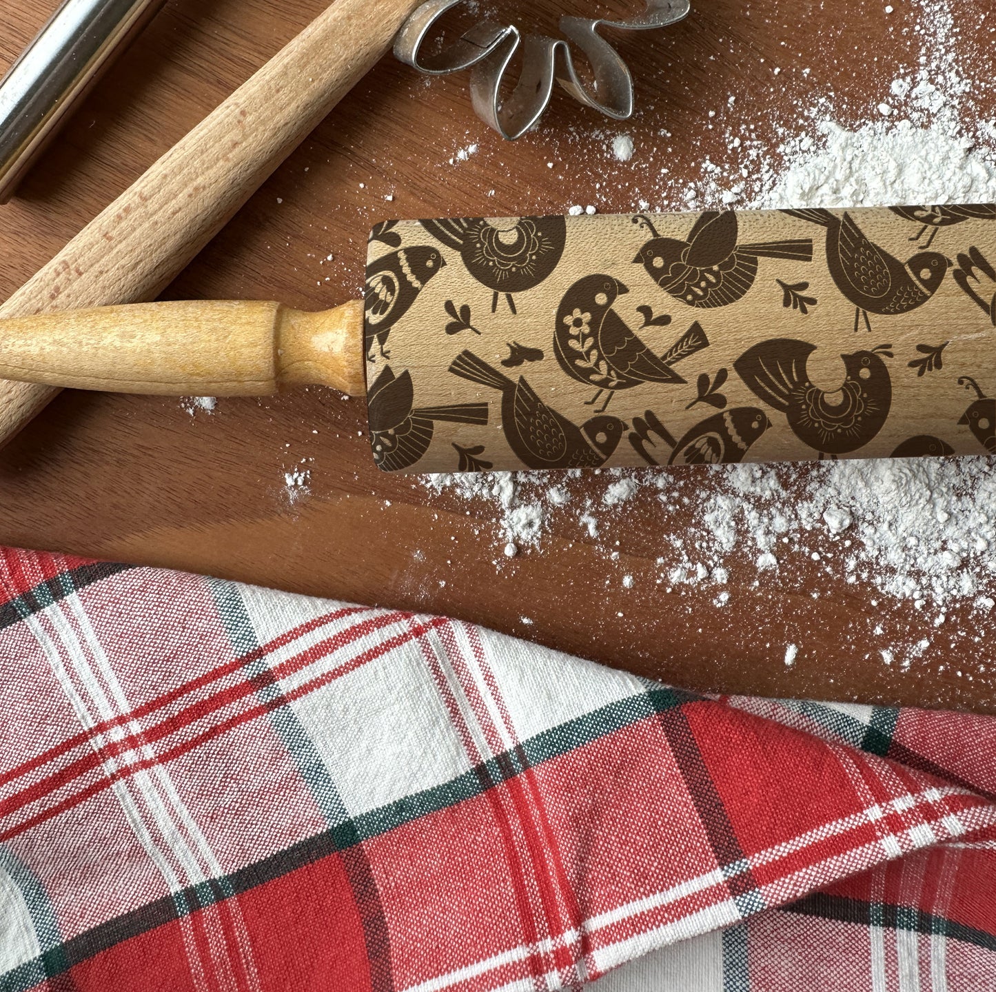 Wooden rolling pin with bird pattern on a wooden surface with flour and plaid fabric.