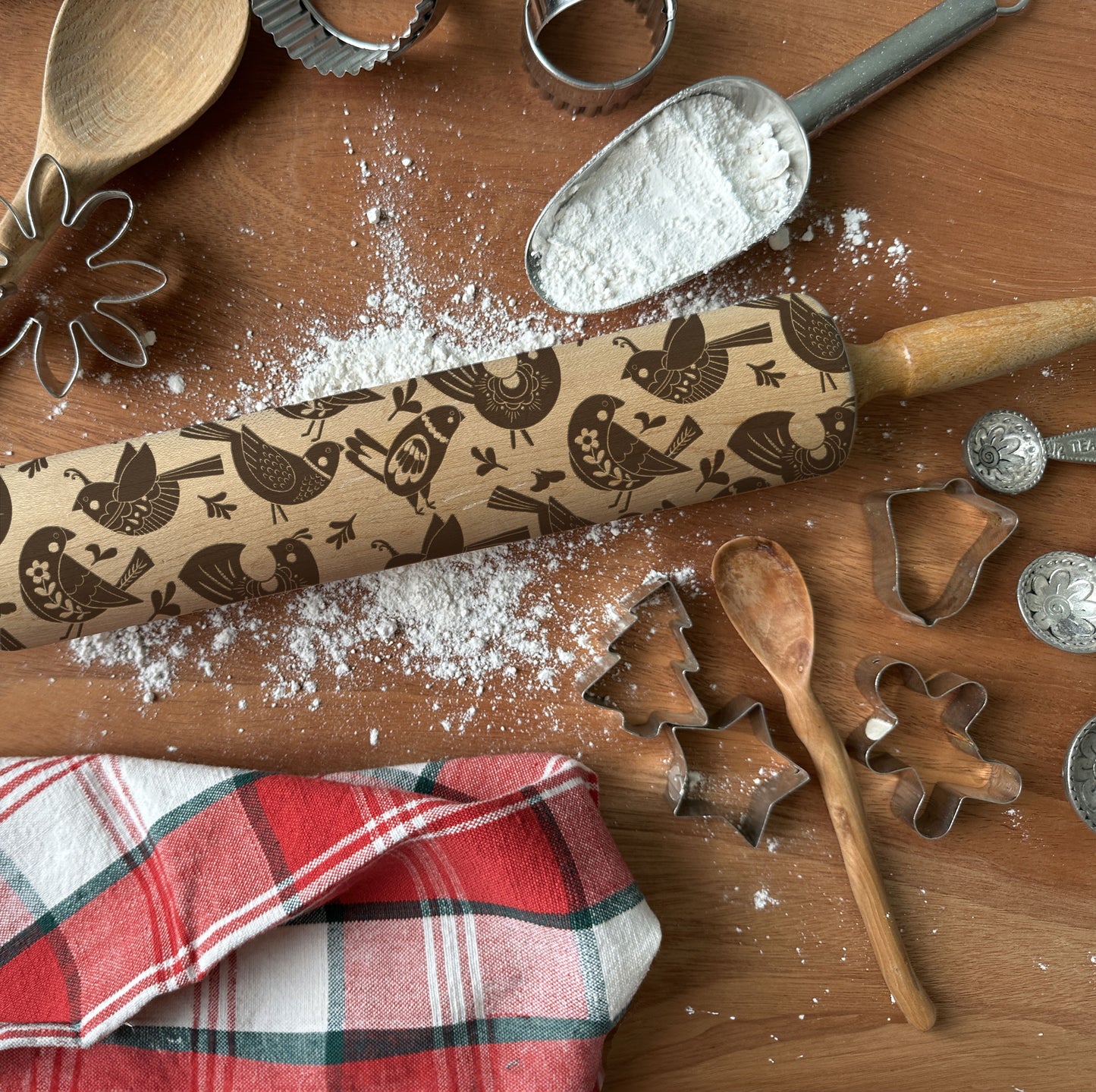 Wooden rolling pin with bird pattern on a wooden surface with flour, cookie cutters, and a red checkered towel.