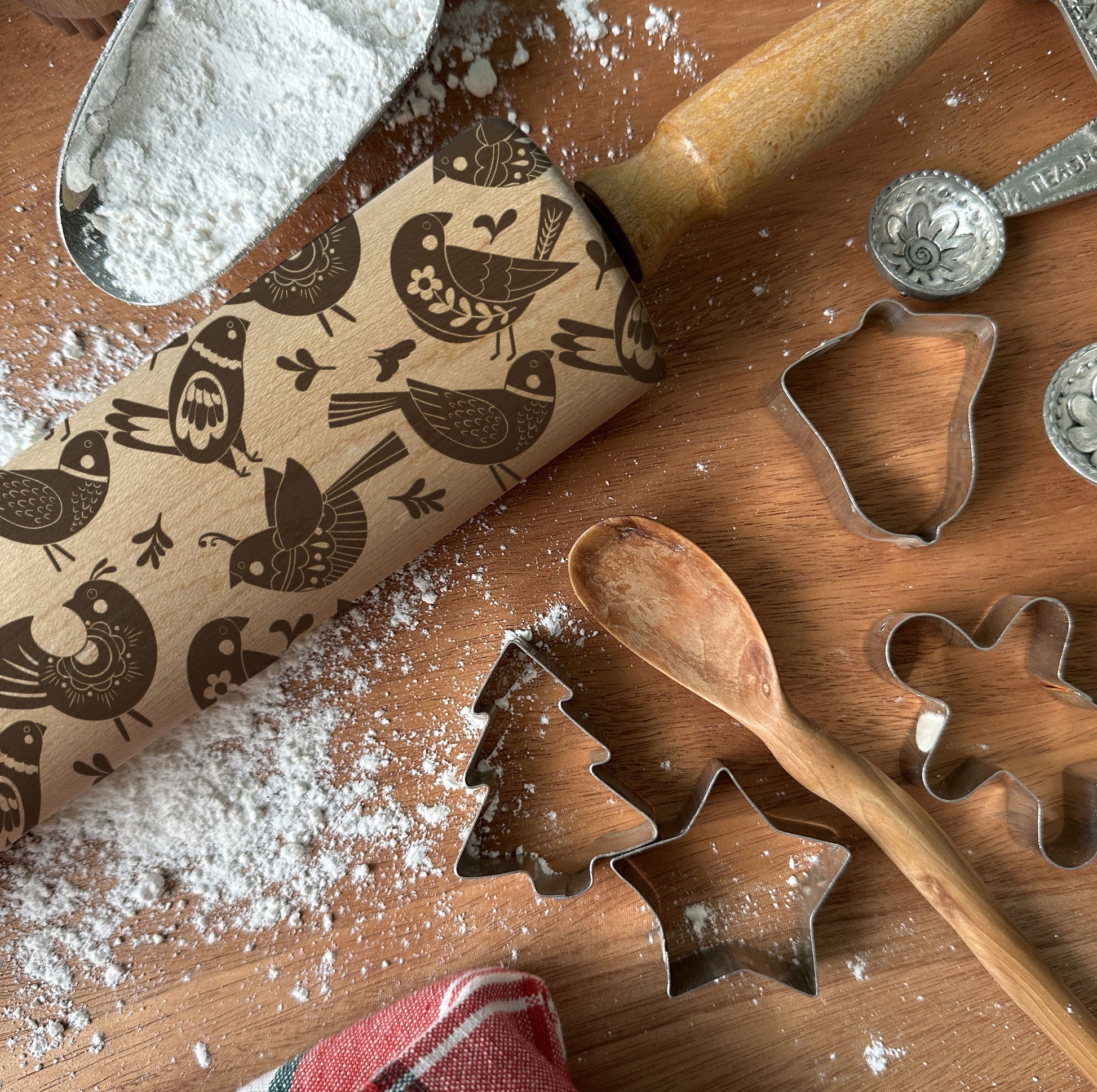 Wooden rolling pin with bird pattern and cookie cutters on a wooden surface with flour.