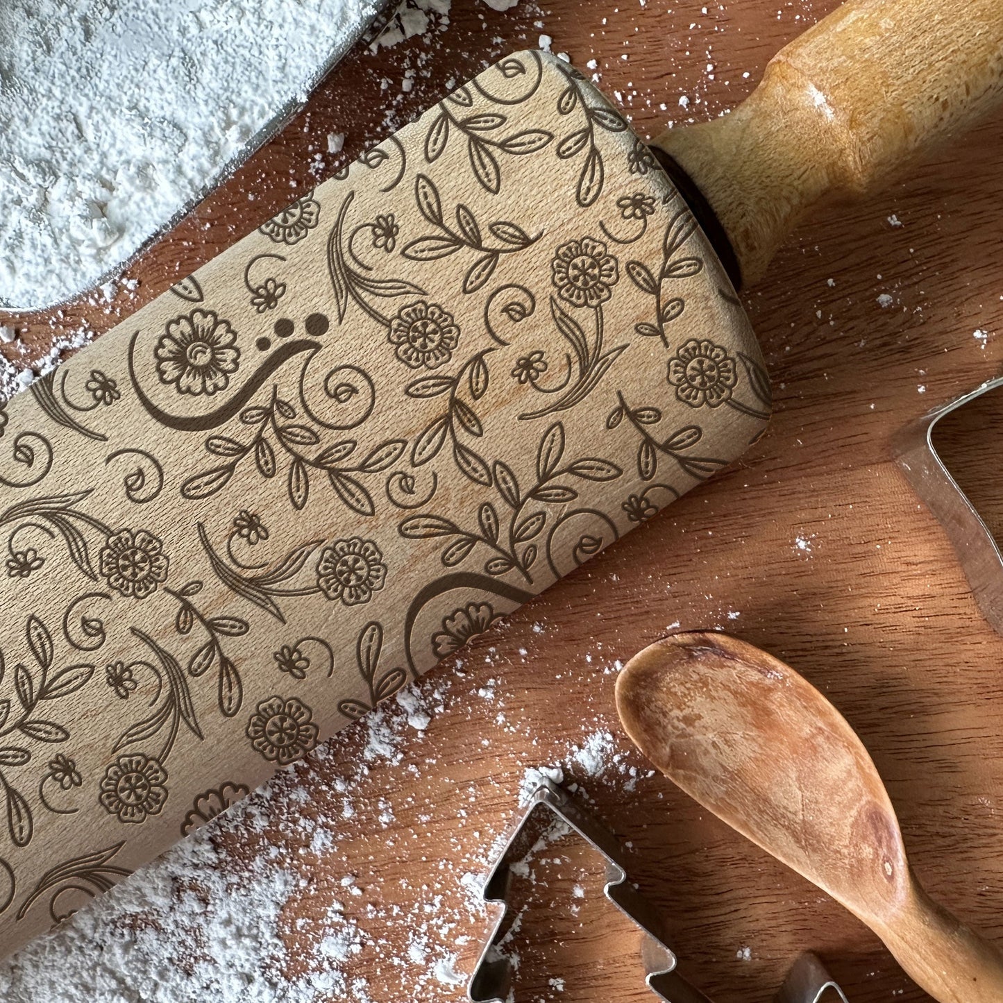 Decorative rolling pin with floral patterns on a wooden surface with flour and cookie cutters.