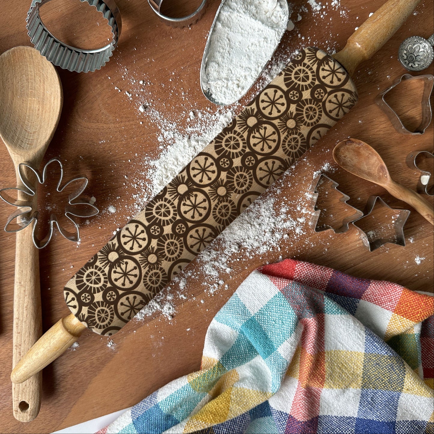 Baking tools including a rolling pin with embossed patterns, wooden spoon, whisk, and flour on a wooden surface.