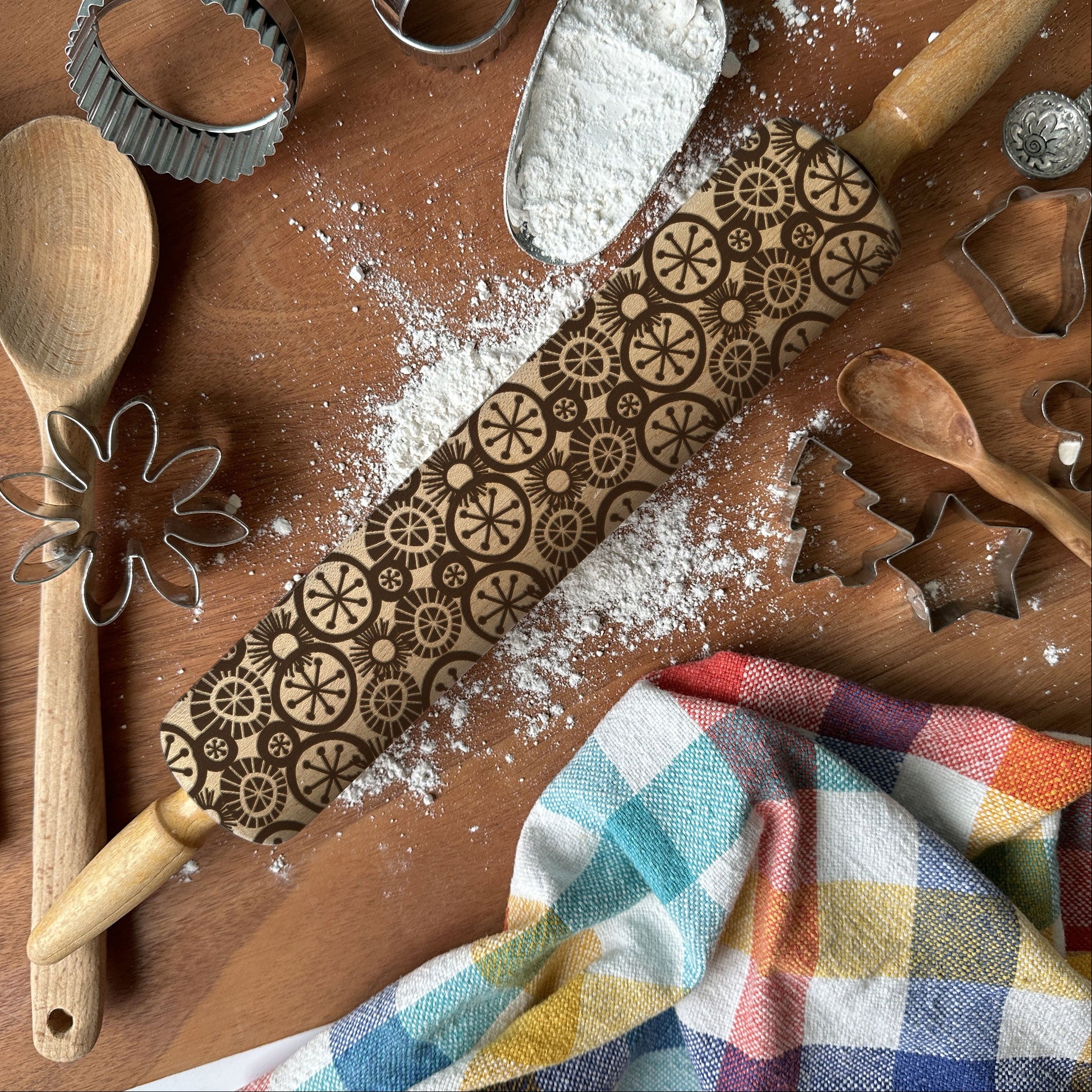 Baking tools including a rolling pin with embossed patterns, wooden spoon, whisk, and flour on a wooden surface.