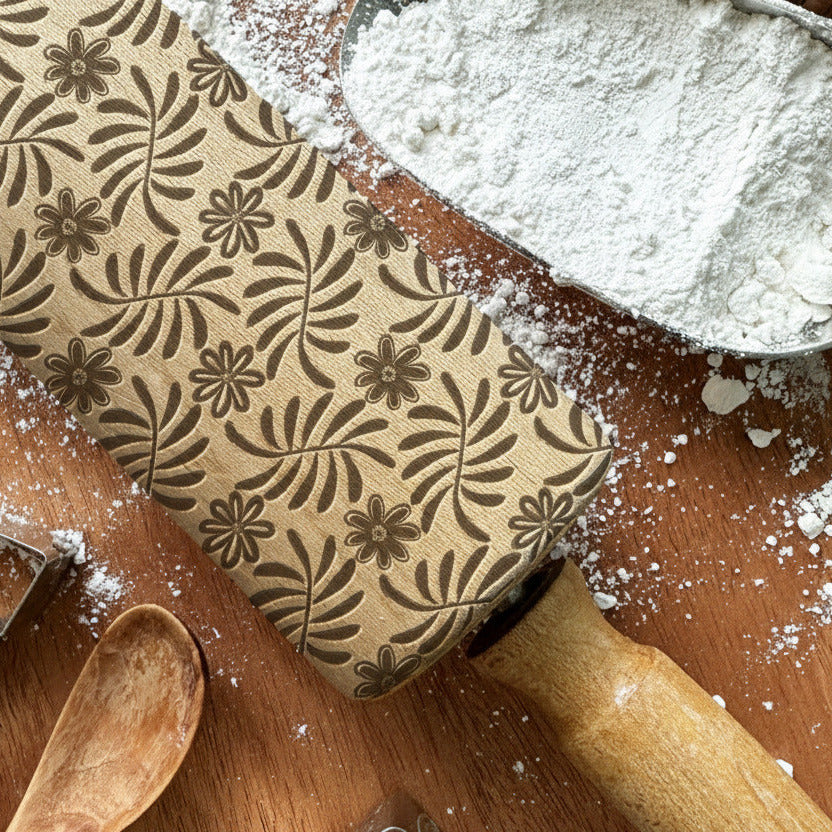 Baking tools including a rolling pin with a floral pattern, cookie cutters, and a wooden spoon on a wooden surface with flour.