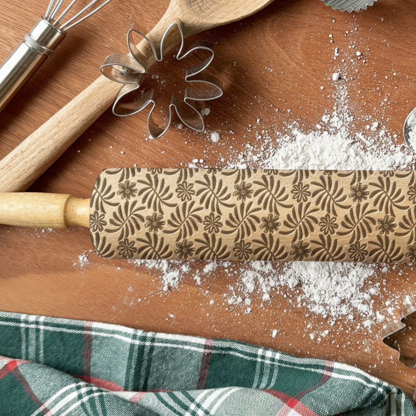 Rolling pin with embossing pattern on a wooden surface with flour, plaid fabric, and cookie cutters.