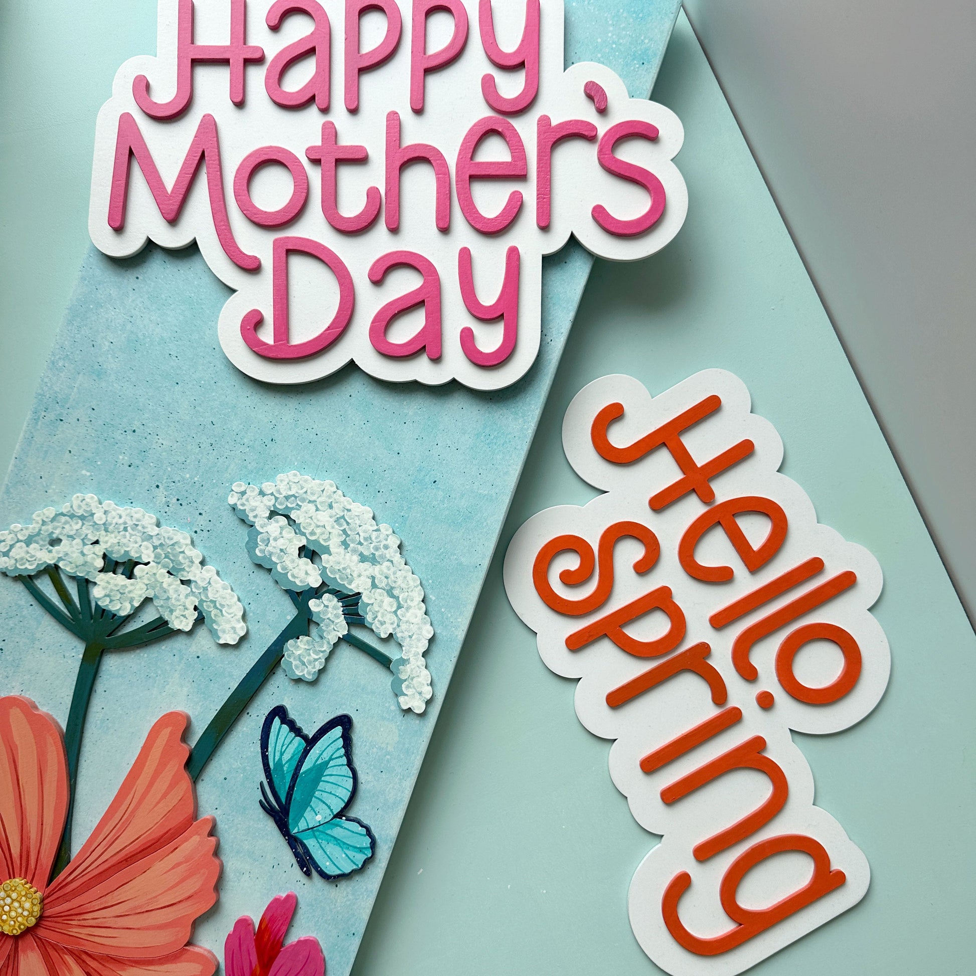 Sign with flowers and 'Happy Mother's Day' text, featuring an optional 'Hello Spring' saying on a light blue background.