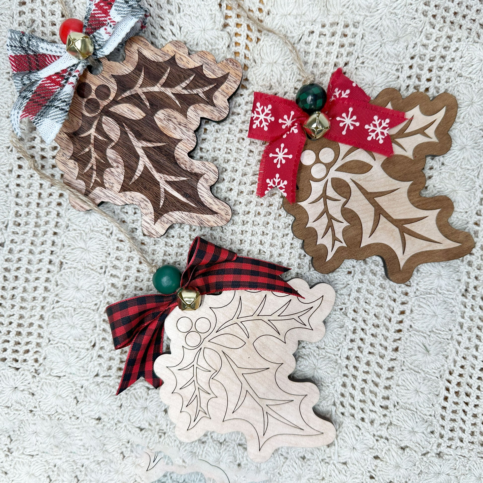 Three wooden Christmas ornaments shaped like holly leaves with ribbons on a textured fabric background.