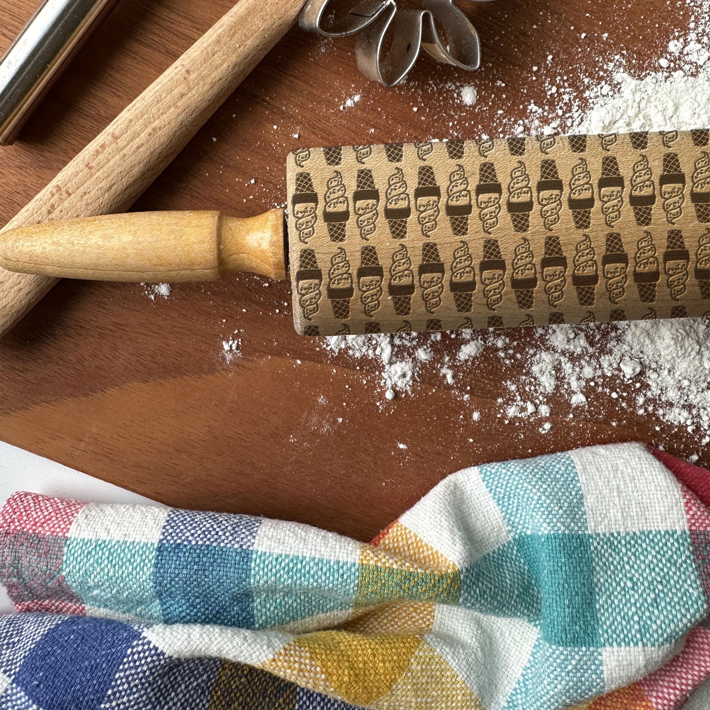 Decorative rolling pin with ice cream cone pattern on a wooden surface with flour and a checkered cloth.