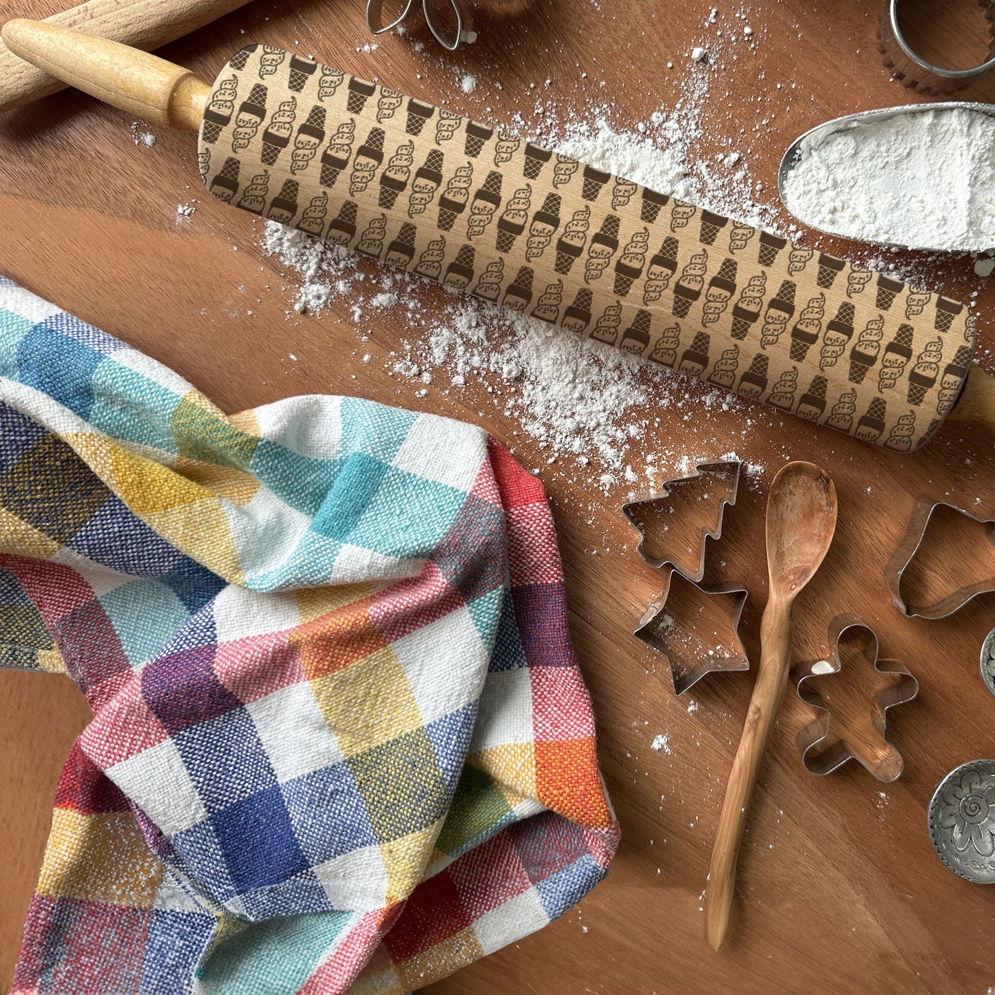 Baking tools including a patterned rolling pin, cookie cutters, and a wooden spoon on a wooden surface with flour.