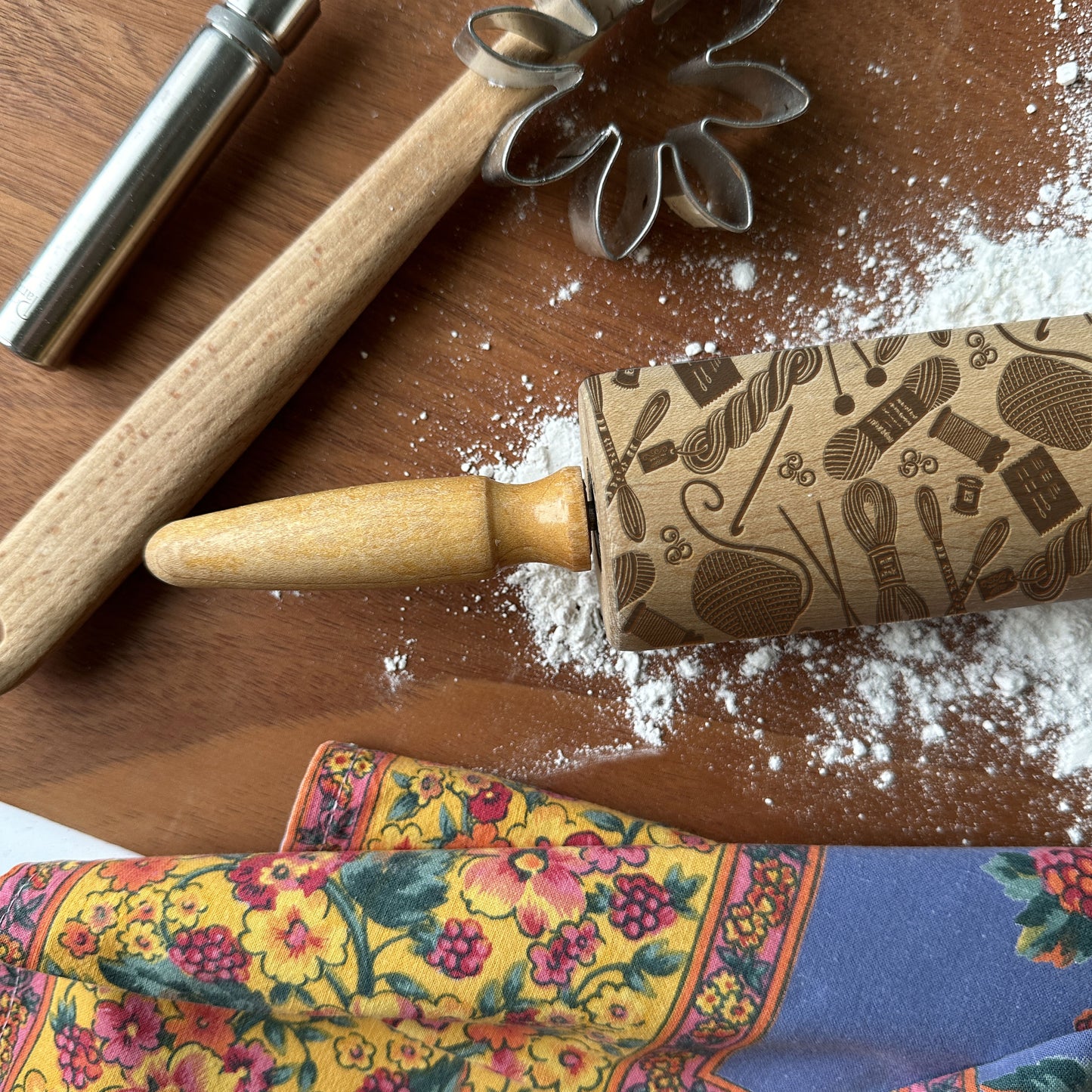 Wooden rolling pin with patterned handle on a wooden surface with flour and cookie cutters.