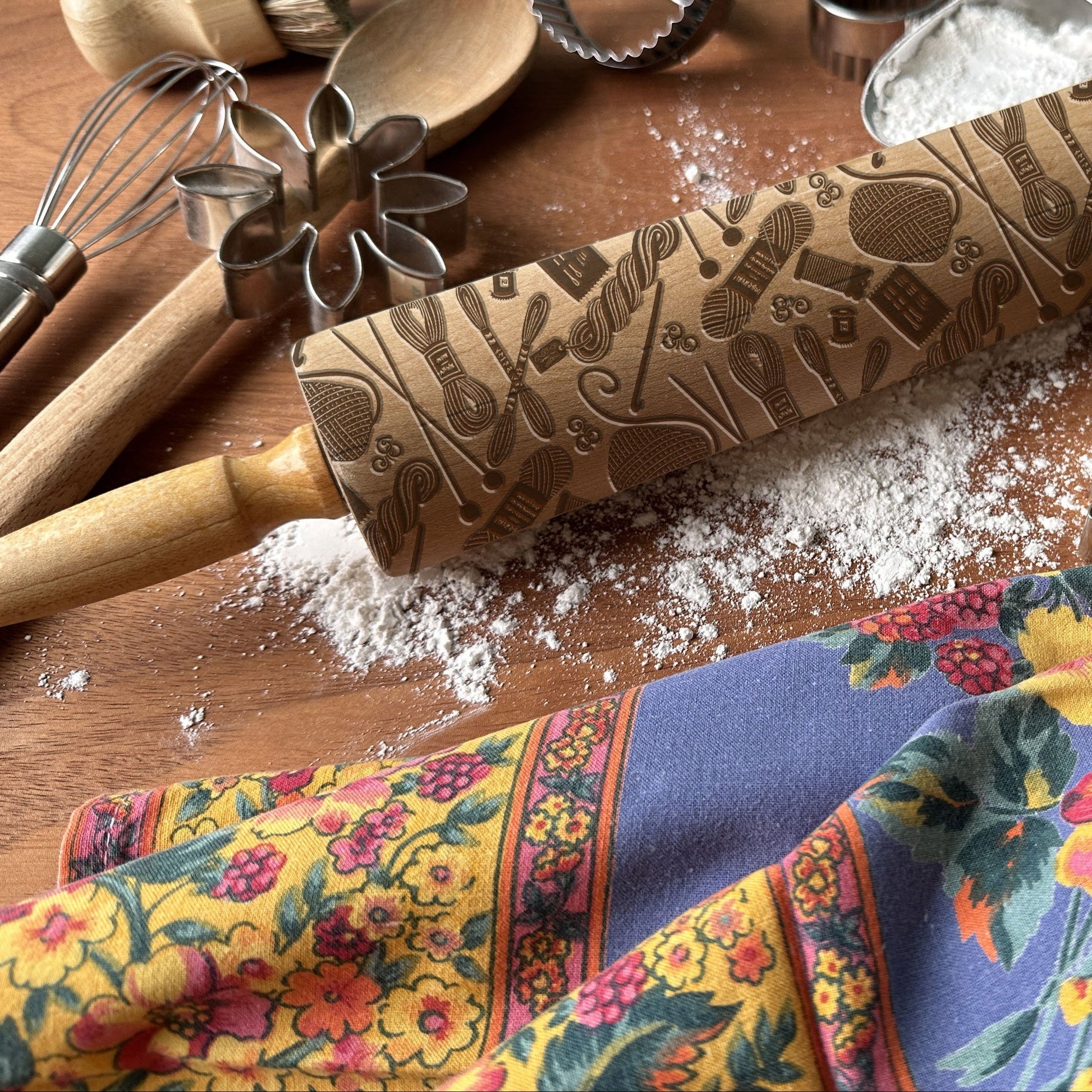 Decorative rolling pin with embossed patterns on a wooden surface with kitchen utensils and a colorful towel.