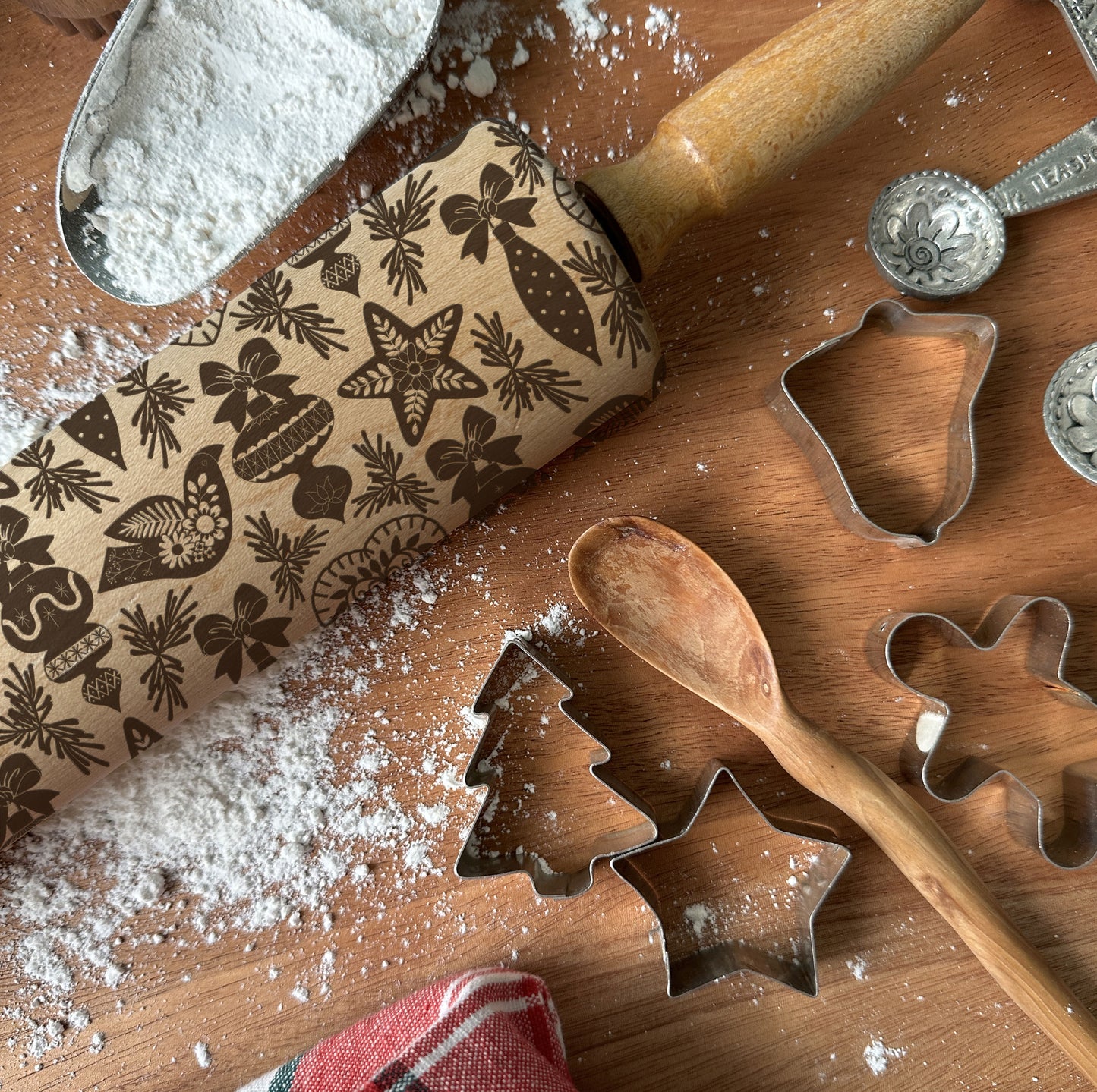 Wooden rolling pin with Christmas patterns, cookie cutters, and flour on a wooden surface.
