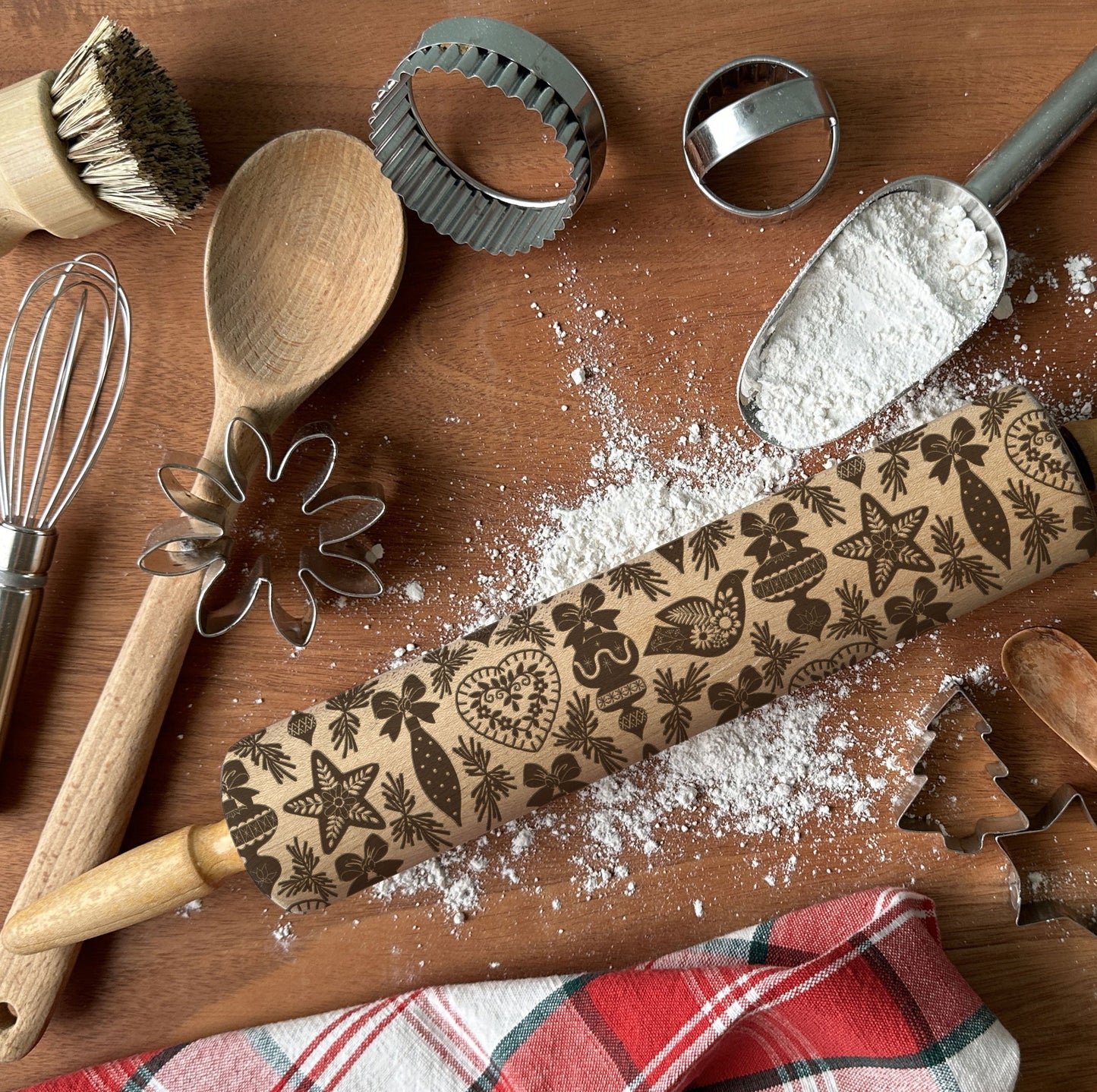 Baking tools including a rolling pin with embossed designs, wooden spoon, whisk, and cookie cutters on a wooden surface with flour.